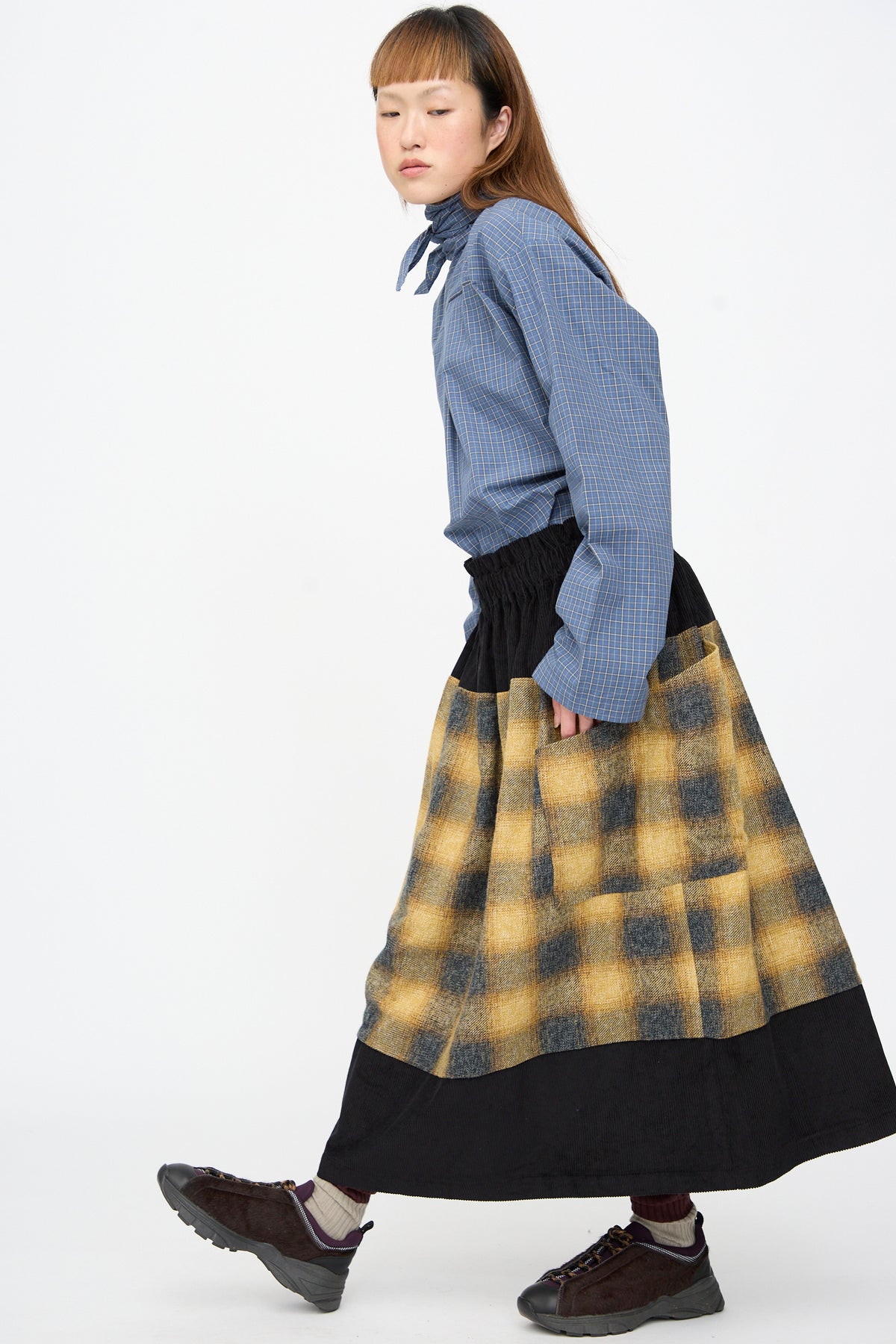 A person wearing a blue checkered shirt and the SC103 Cotton Flannel Rubric Skirt in Hearth, an oversized plaid maxi skirt with black panels, paired with dark sneakers.