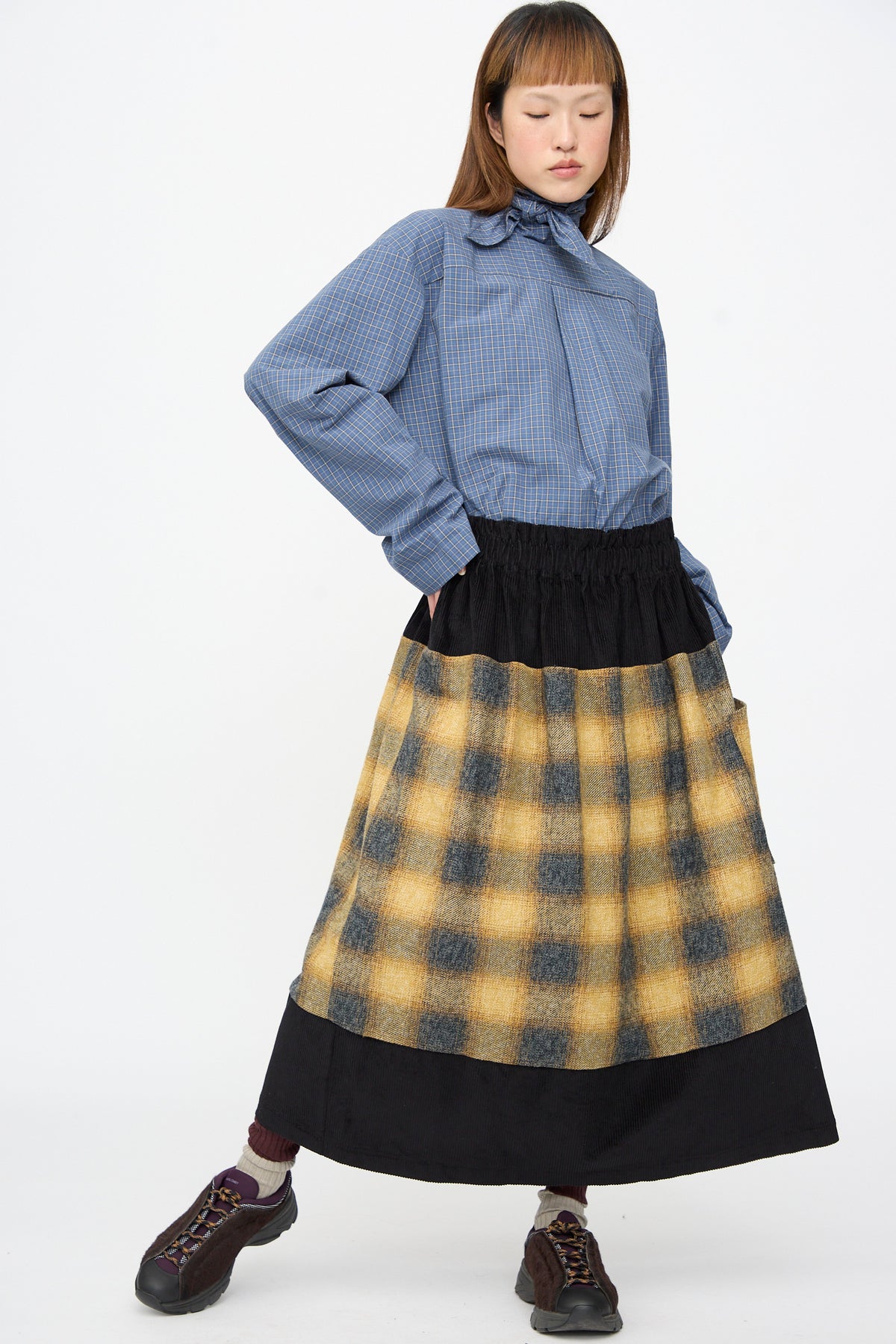 A person wears a blue plaid collared shirt and an SC103 Cotton Flannel Rubric Skirt in Hearth, featuring black panels and yellow and black plaid middle section.