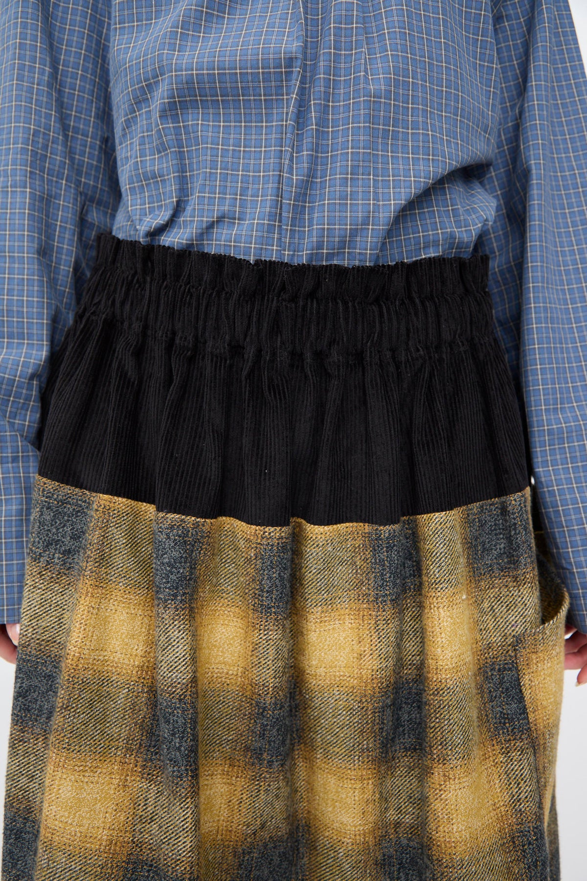 A SC103 Cotton Flannel Rubric Skirt in Hearth, featuring a midi-length, black elastic waistband and a yellow and grey plaid lower panel, worn with a blue plaid button-down shirt.