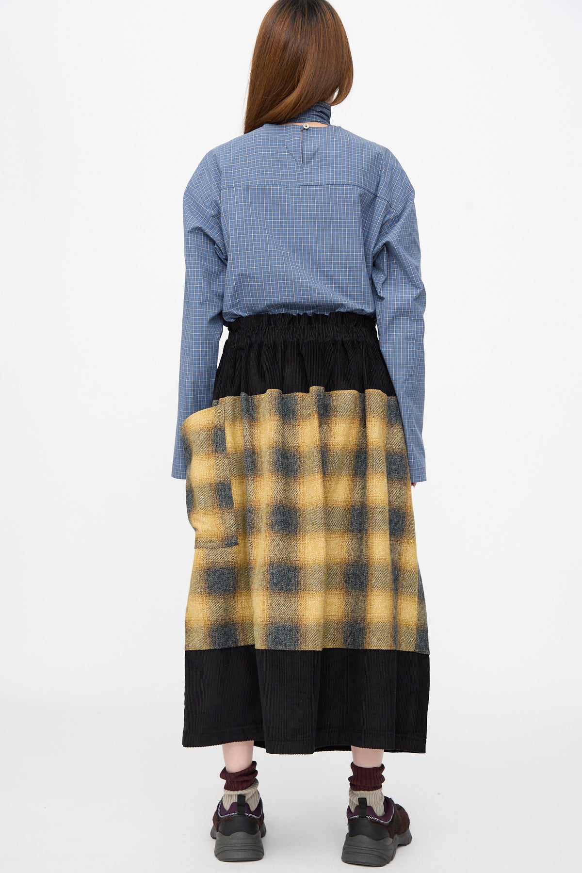 A person seen from behind wears a blue button-down shirt and a Cotton Flannel Rubric Skirt in Hearth with yellow plaid and solid black panels, paired with dark sneakers.