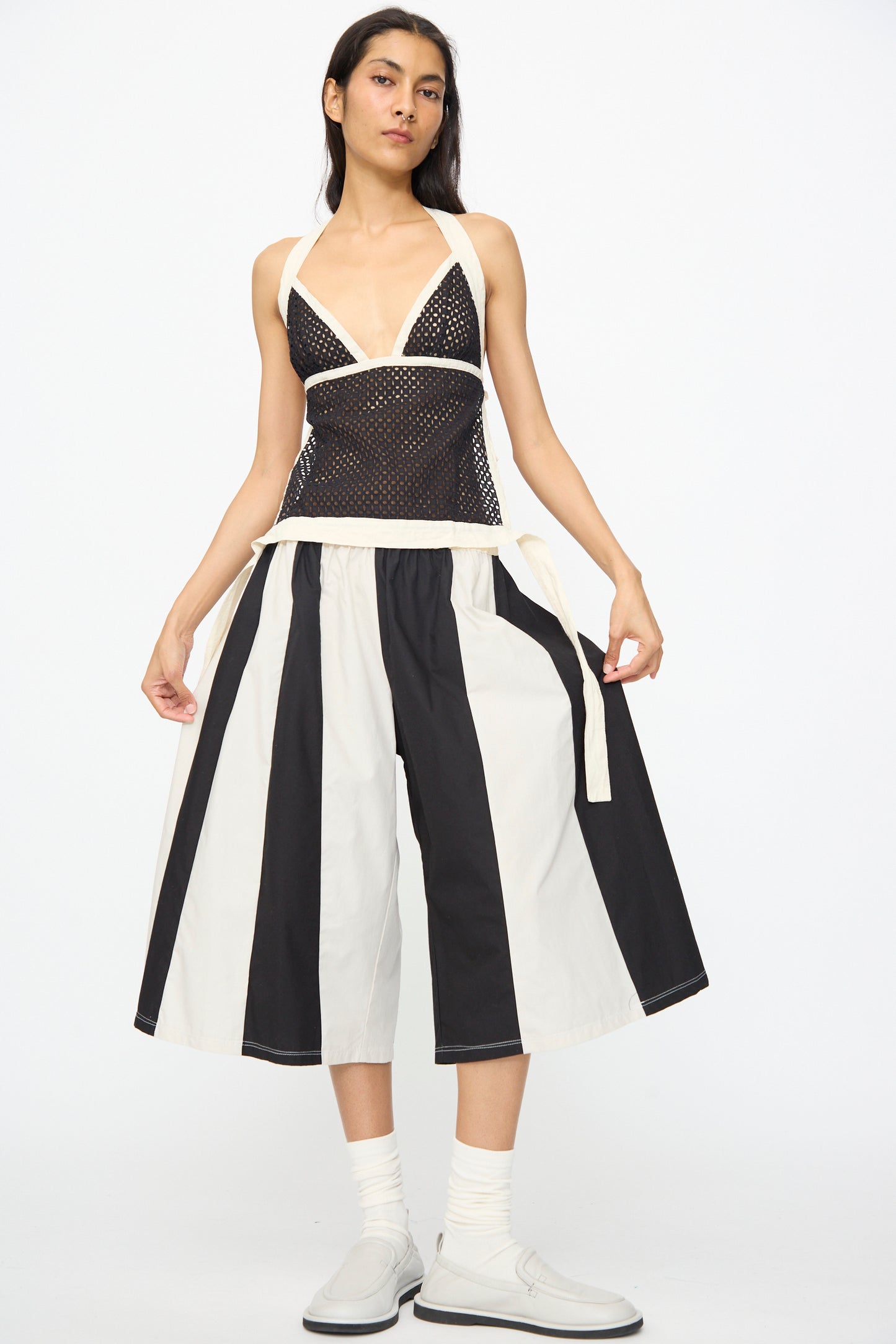 A person in SC103’s Cotton Poplin Pennant Shorts in Helix, paired with a black mesh top with white straps and white socks and shoes, stands against a plain white background.