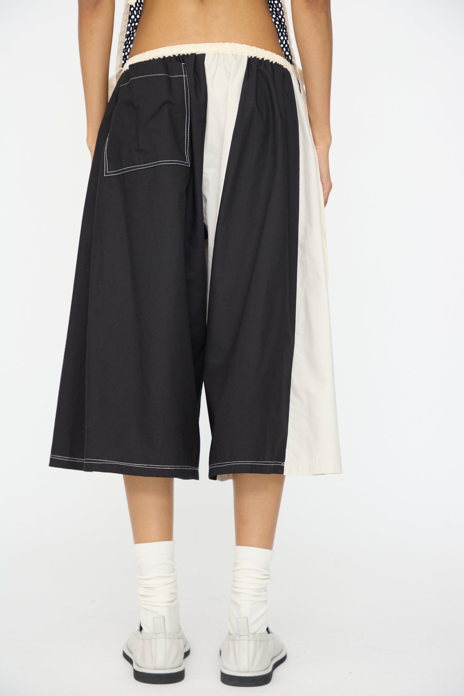 A person wearing SC103 Cotton Poplin Pennant Shorts in Helix, featuring a relaxed fit and single back pocket, paired with white socks and sandals, is shown from the back against a plain background.