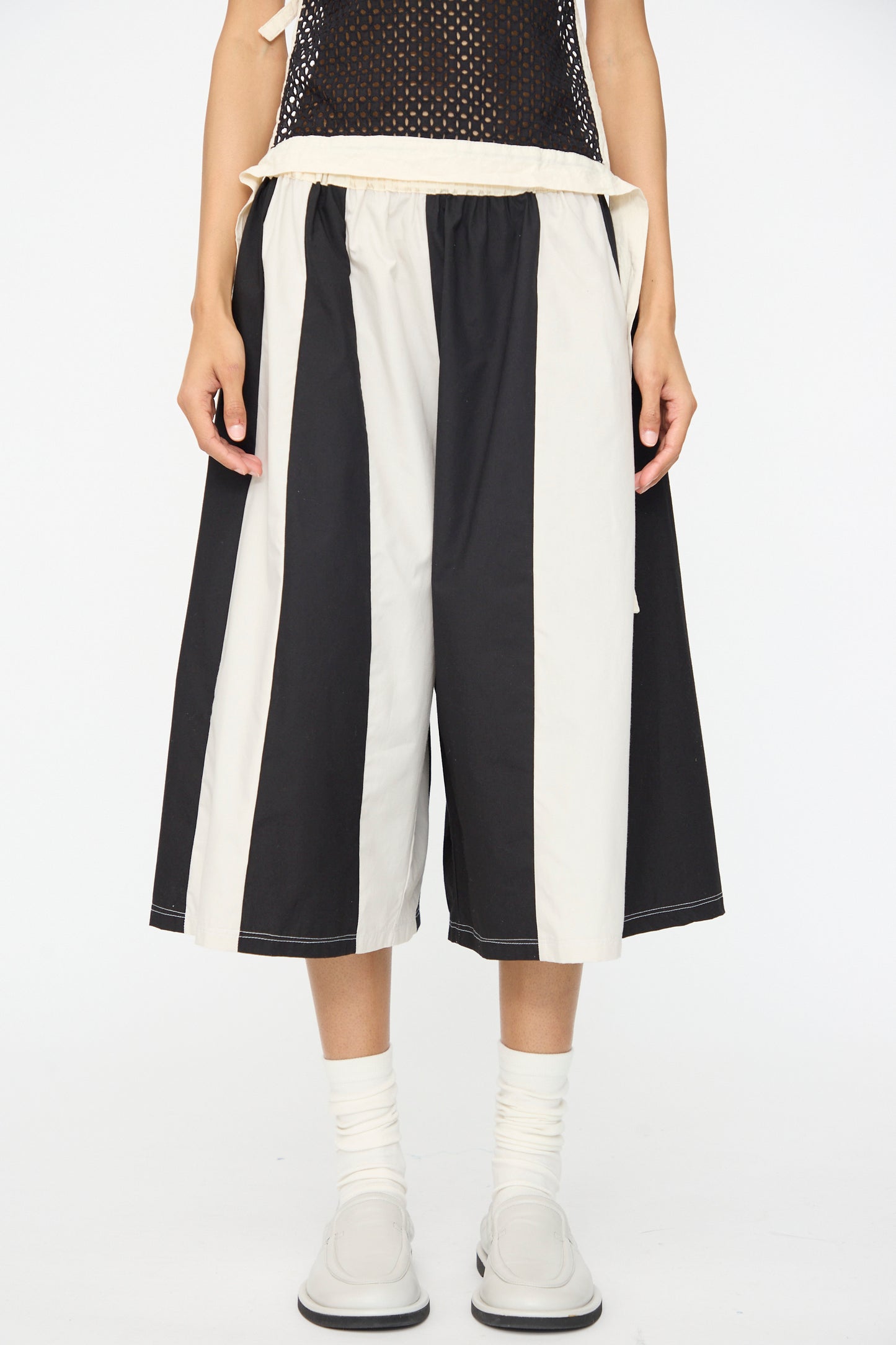 A person wearing SC103's Cotton Poplin Pennant Shorts in Helix, featuring relaxed fit, knee-length black-and-white stripes, paired with white socks and light-colored shoes.