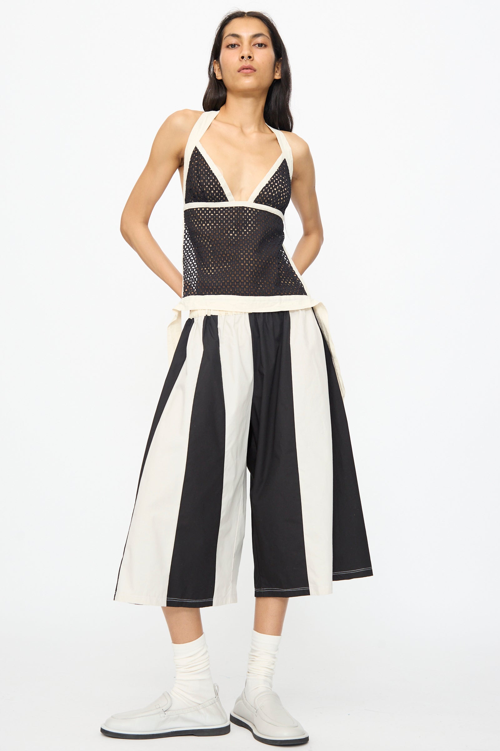 A woman wears SC103 Cotton Poplin Pennant Shorts in Helix—black-and-white striped, relaxed-fit cotton—paired with a black mesh deep v-neck top, white shoes, and socks.