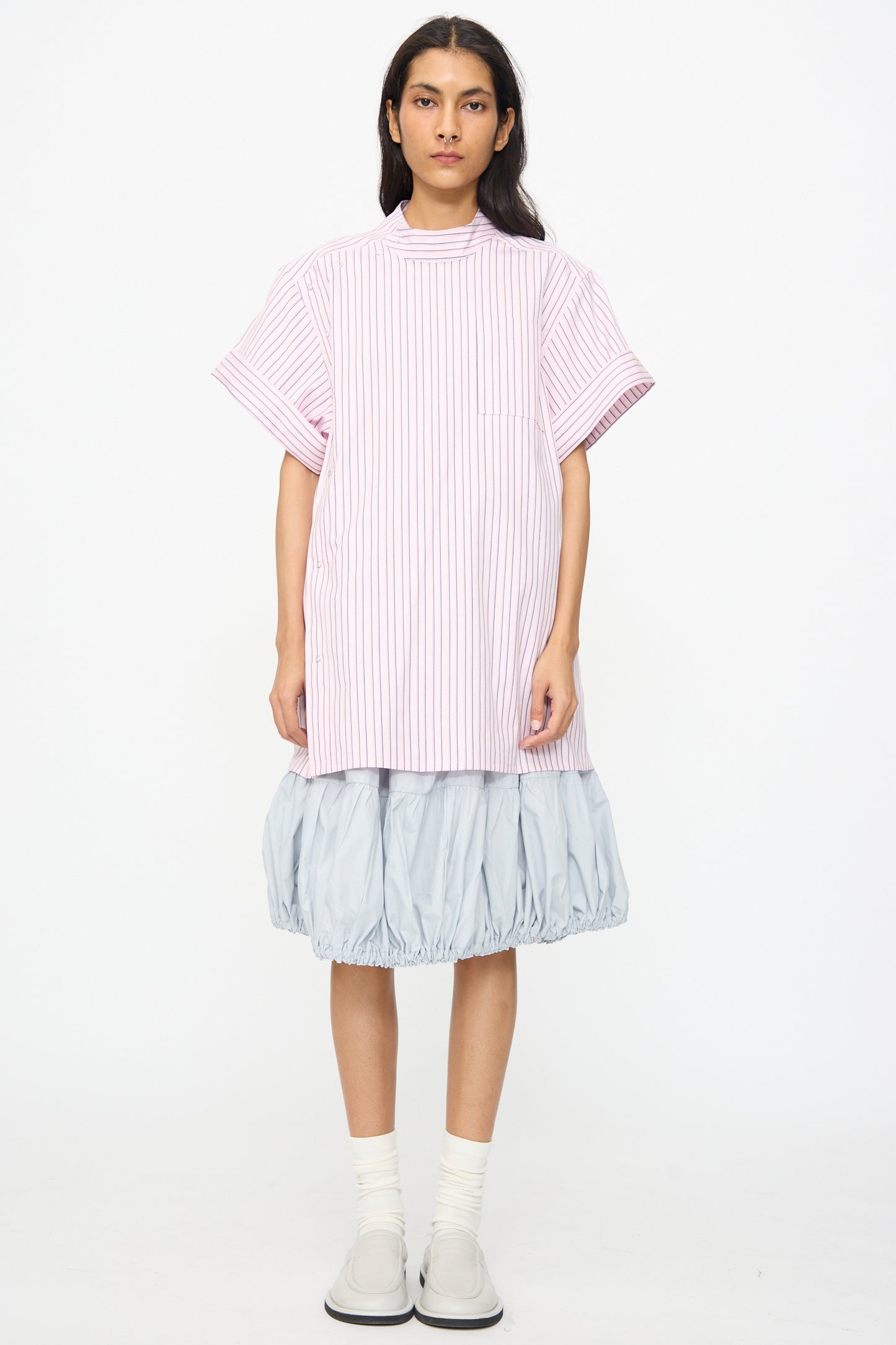 A person stands against a white background wearing a pink striped short-sleeve shirt and the SC103 Cotton Poplin Ramble Skirt in Sonic, paired with white socks and light gray shoes.