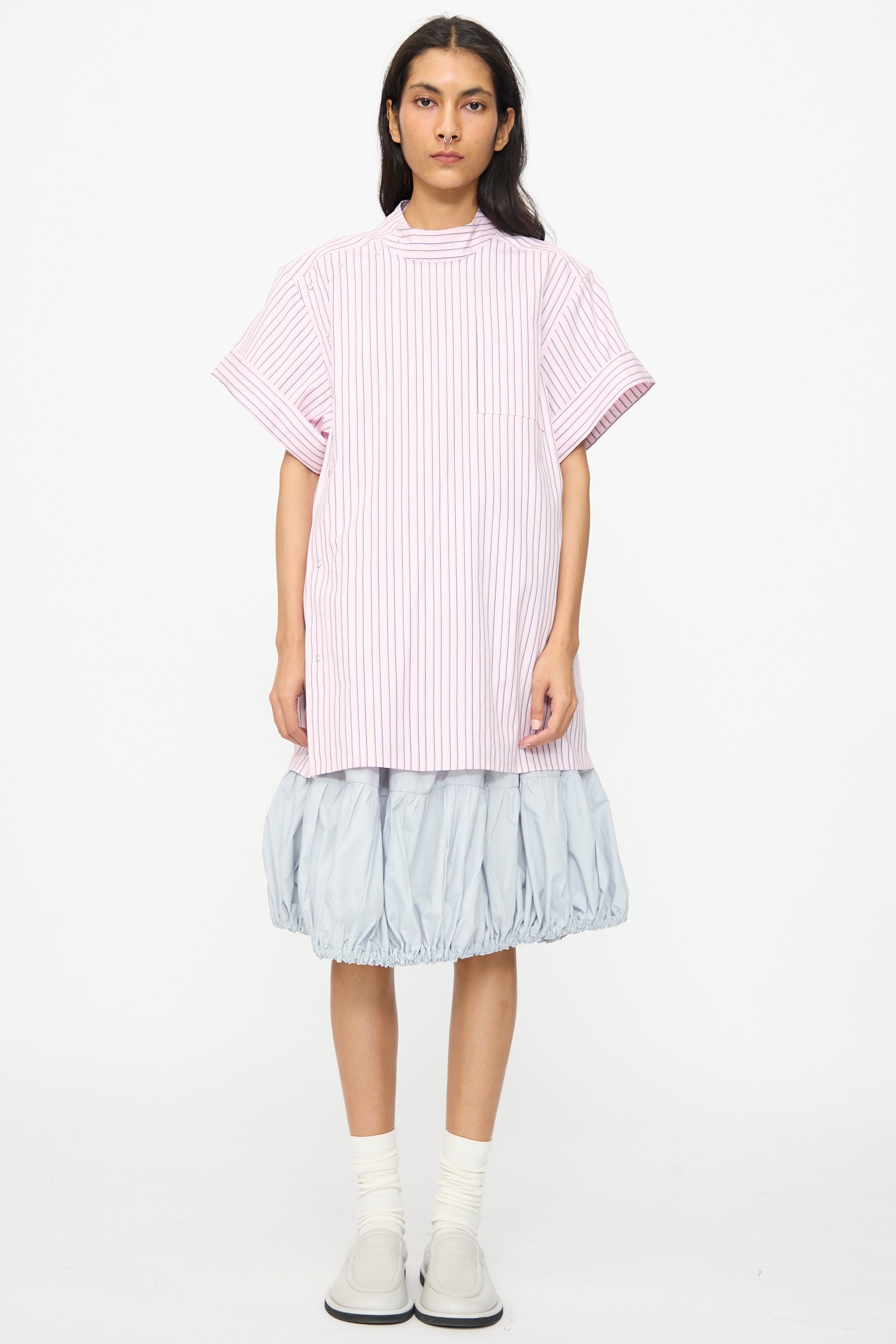 A person stands against a white background wearing a pink striped short-sleeve shirt and the SC103 Cotton Poplin Ramble Skirt in Sonic, paired with white socks and light gray shoes.