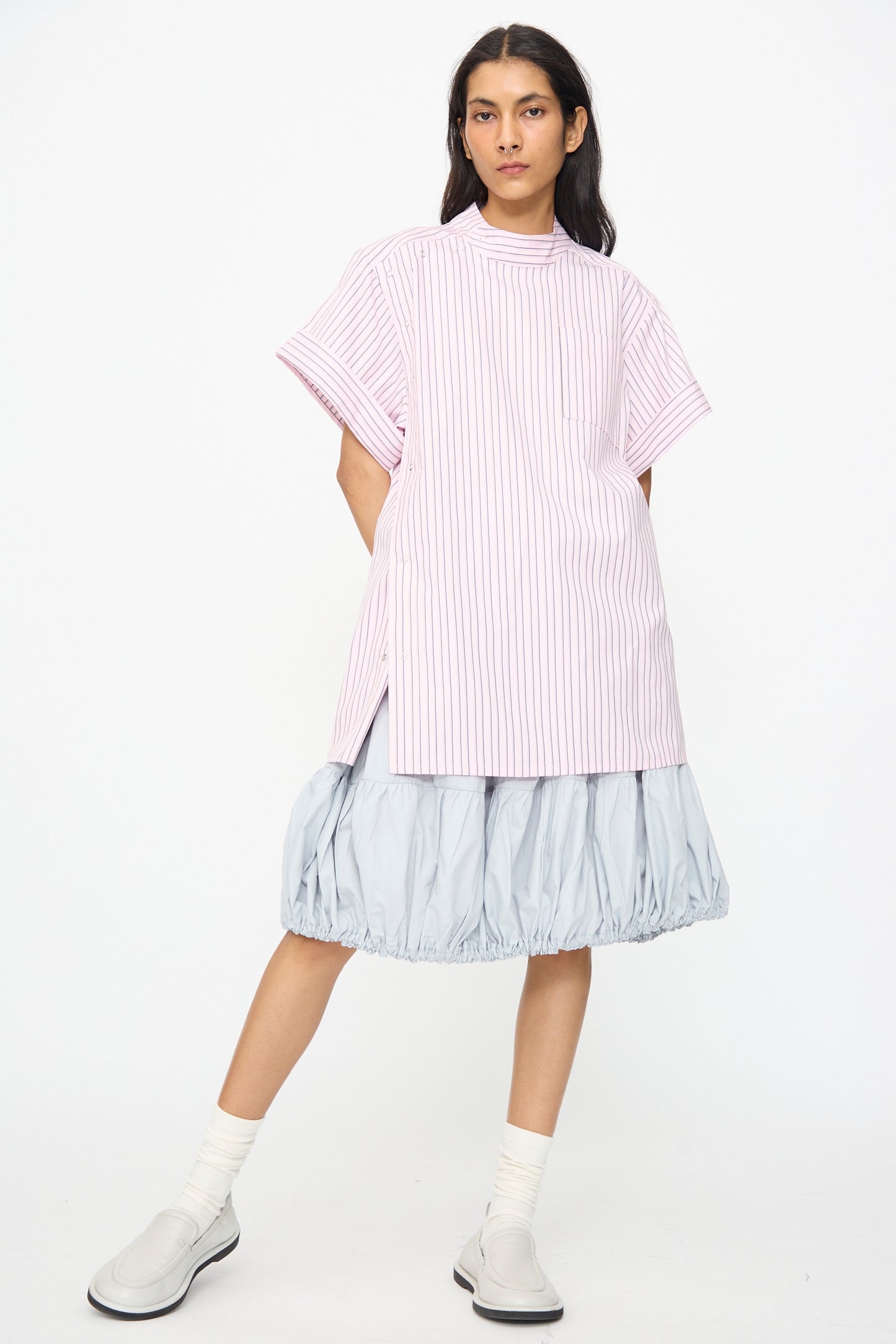 Person standing against a white background wearing a pink-and-white striped oversized shirt, the SC103 Cotton Poplin Ramble Skirt in Sonic, white socks, and light grey shoes.