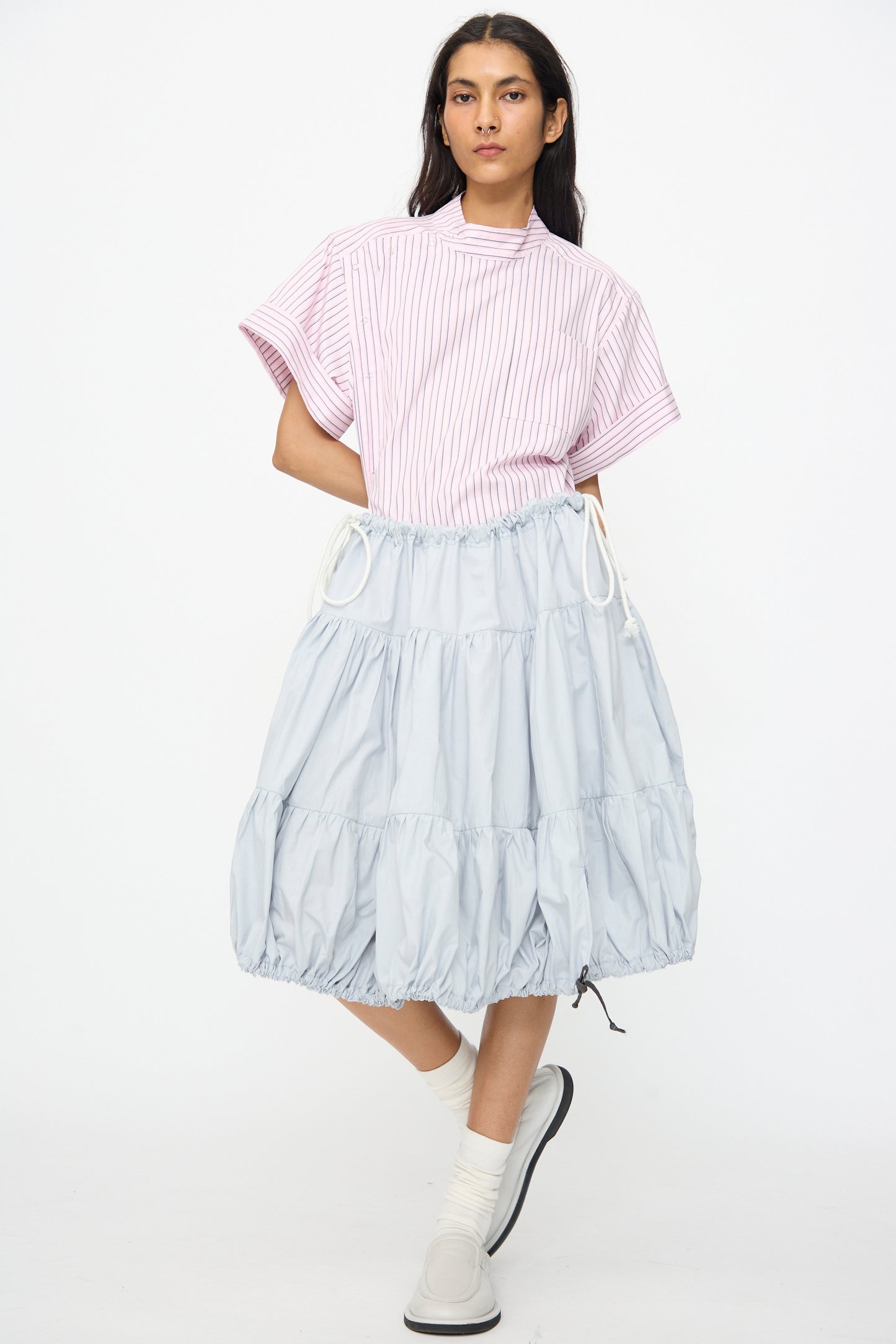 A person stands against a plain white background wearing a pink striped short-sleeve top, the SC103 Cotton Poplin Ramble Skirt in Sonic, white socks, and white slip-on shoes.