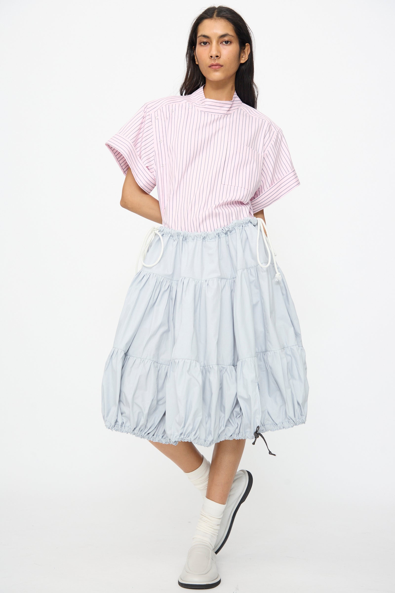 A person stands against a plain white background wearing a pink striped short-sleeve top, the SC103 Cotton Poplin Ramble Skirt in Sonic, white socks, and white slip-on shoes.