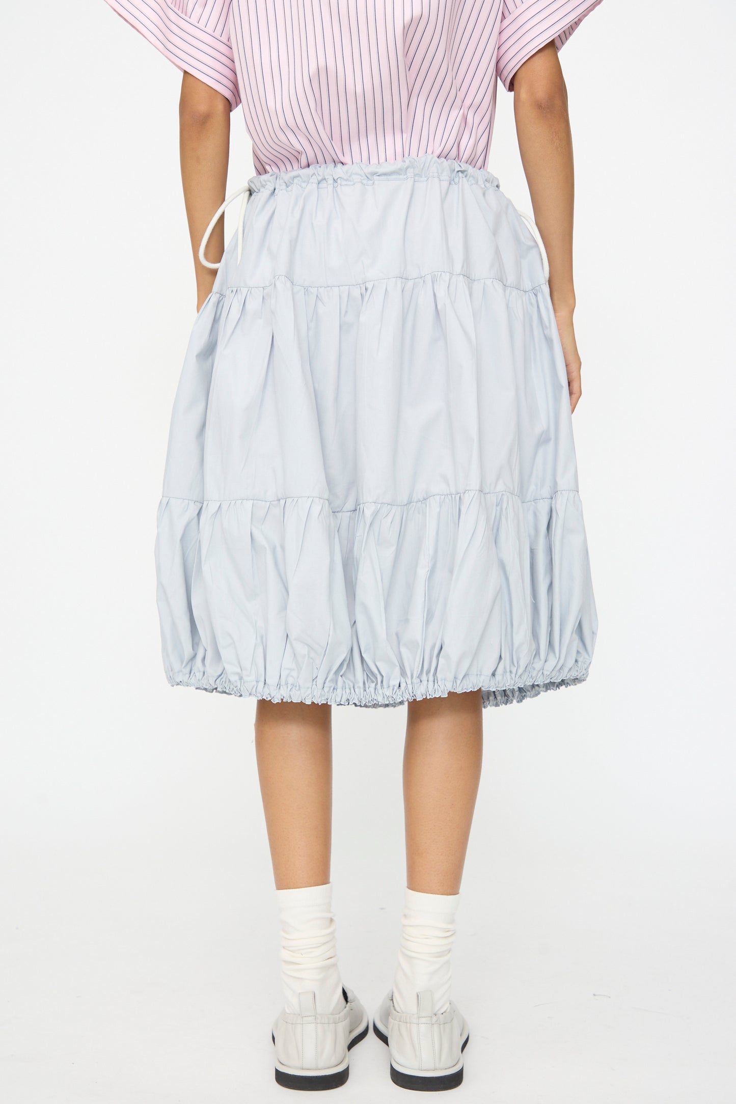 A person is shown from the back wearing the SC103 Cotton Poplin Ramble Skirt in Sonic, which is light blue with tiers and a drawstring waist, paired with white socks, white shoes, and a pink striped top.