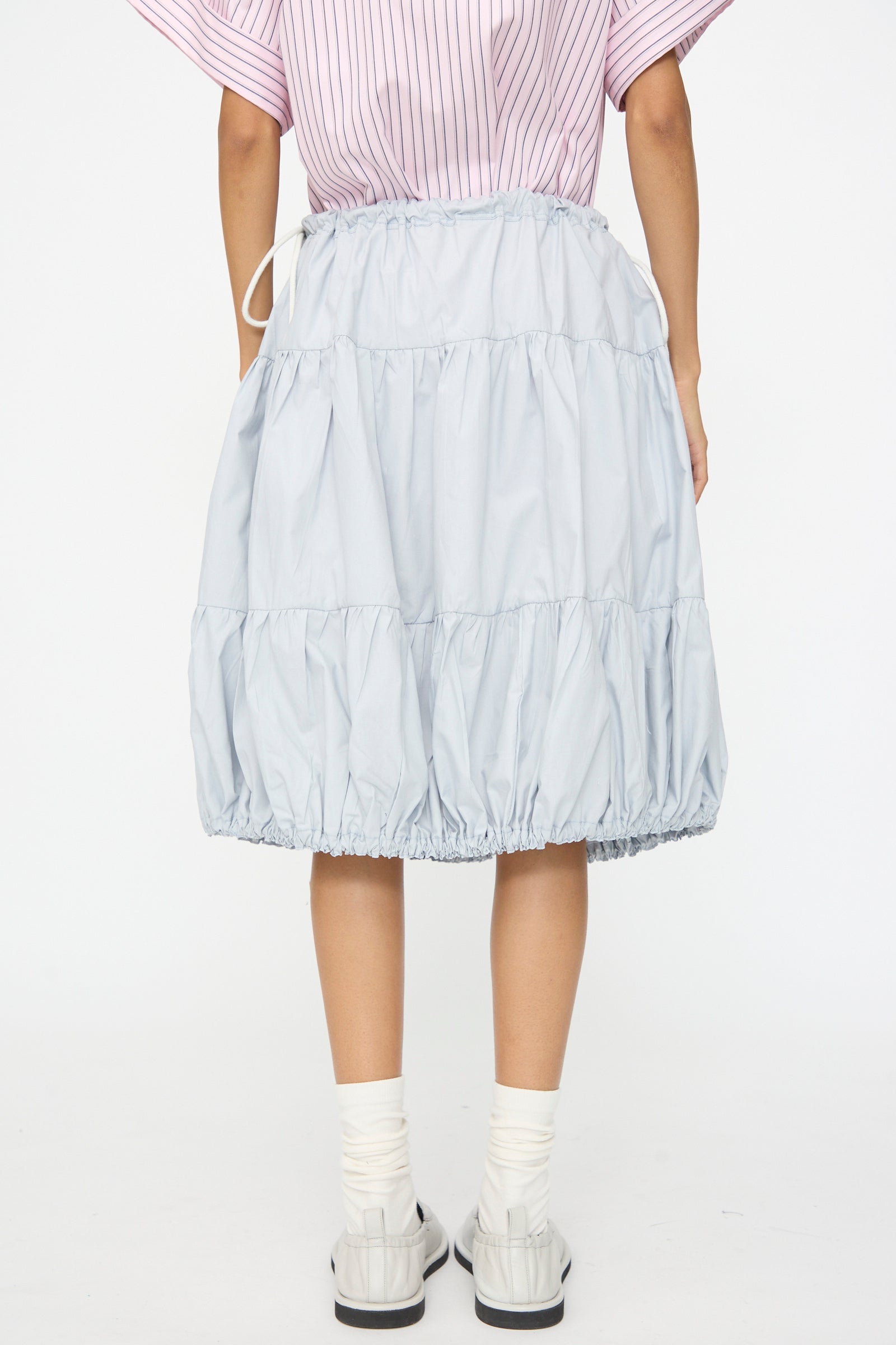 A person is shown from the back wearing the SC103 Cotton Poplin Ramble Skirt in Sonic, which is light blue with tiers and a drawstring waist, paired with white socks, white shoes, and a pink striped top.