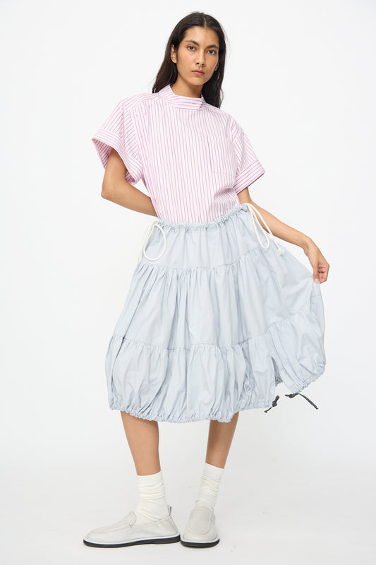 A person stands against a white background wearing a pink striped short-sleeve top, SC103's Cotton Poplin Ramble Skirt in Sonic, white socks, and light-colored shoes.