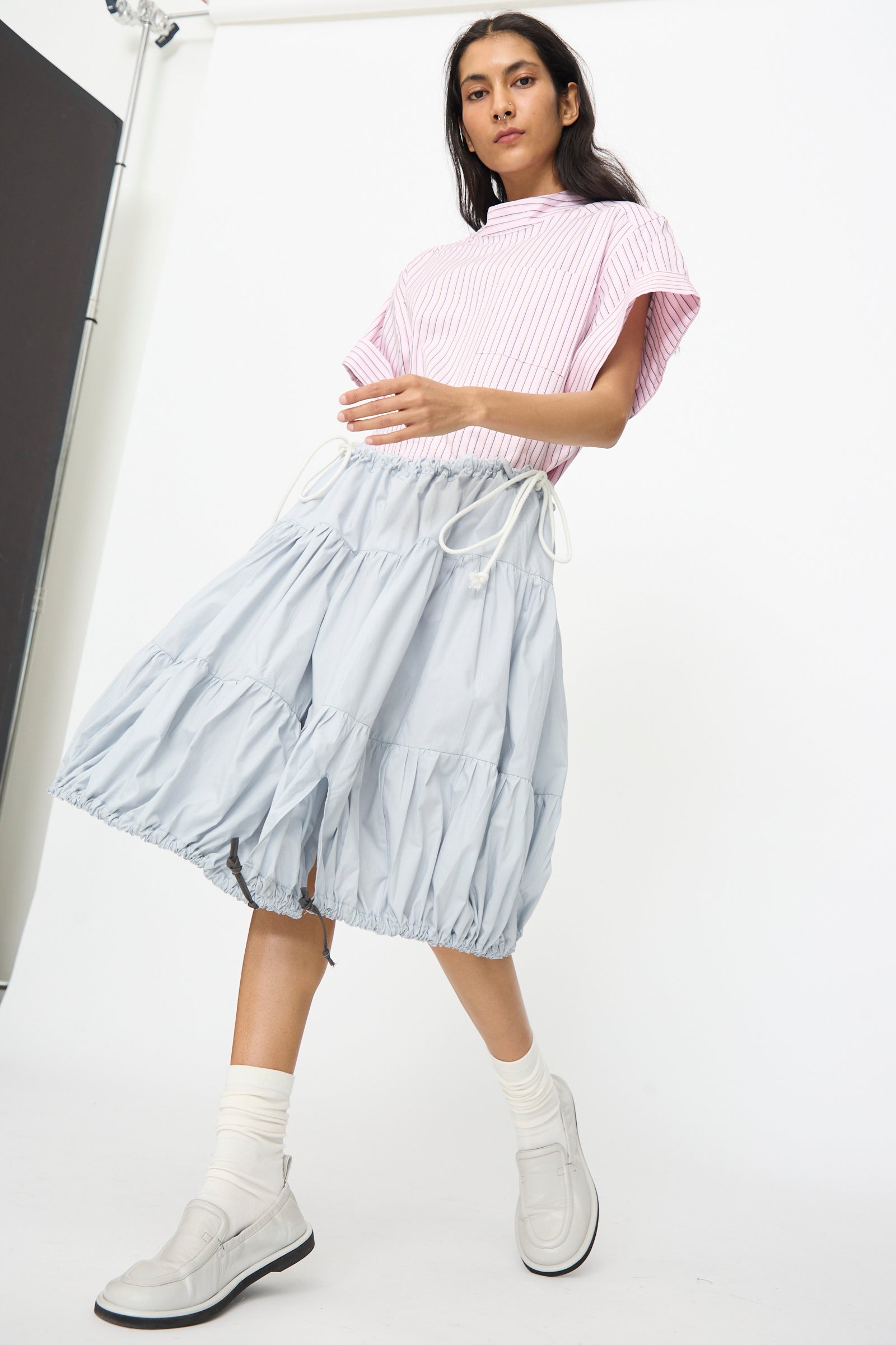 A woman stands against a white background wearing a pink striped top and the SC103 Cotton Poplin Ramble Skirt in Sonic, styled with white socks and white boots.