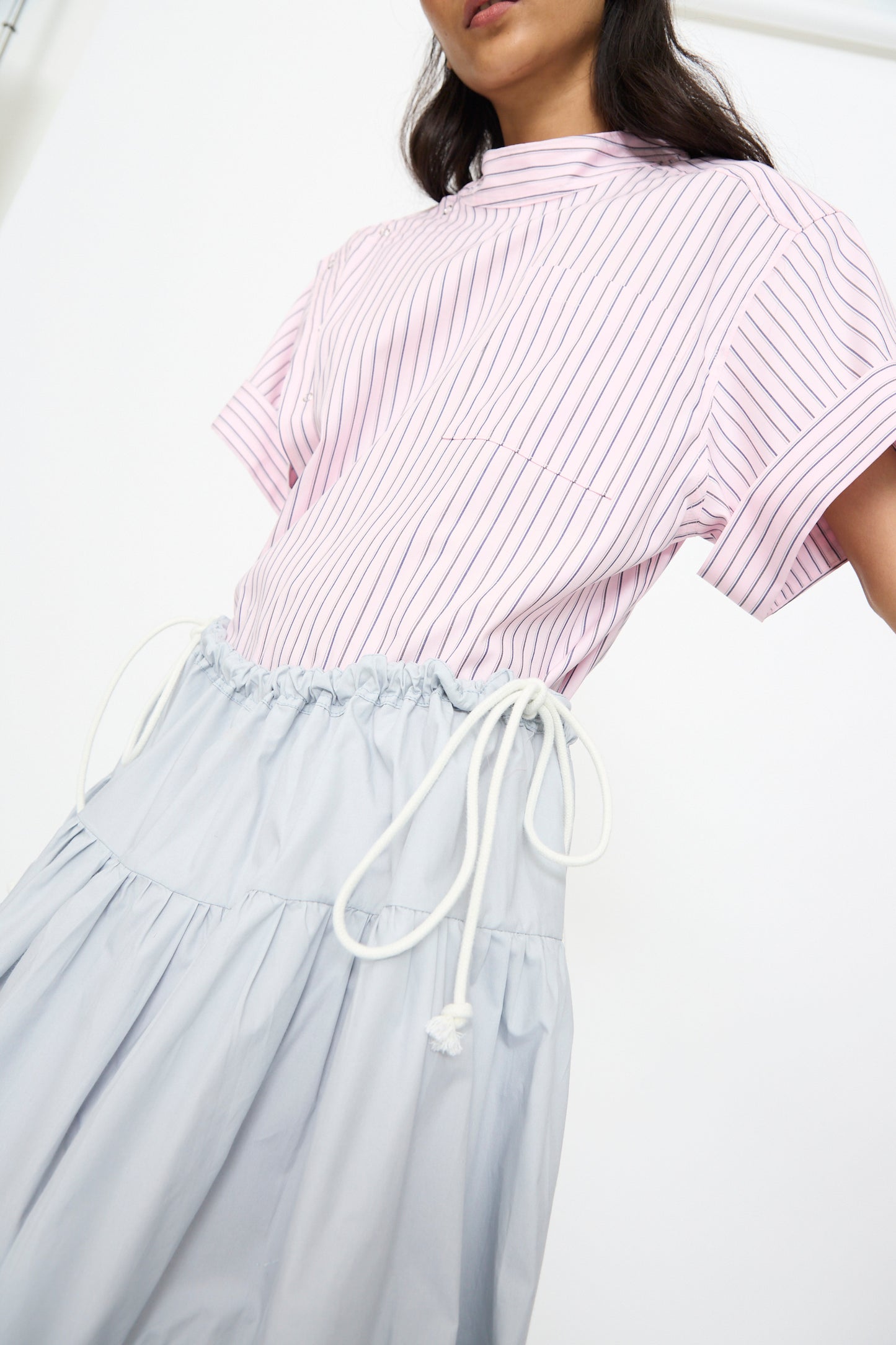 A person wearing a light pink, short-sleeved, pinstriped top and the SC103 Cotton Poplin Ramble Skirt in Sonic, shown from shoulders to knees against a plain background.