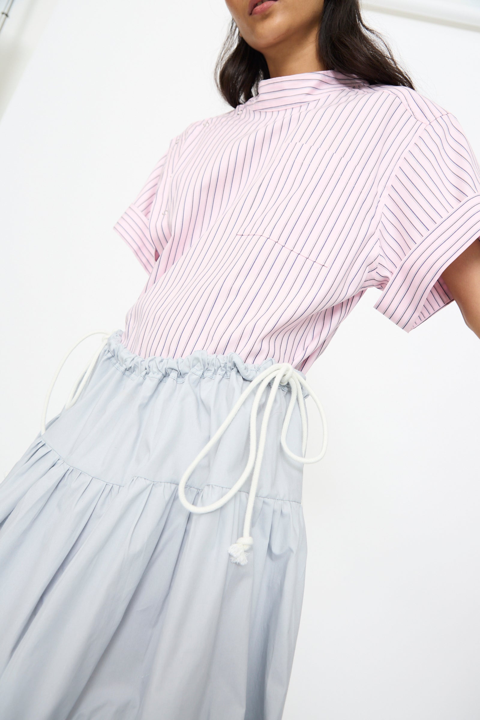 A person wearing a light pink, short-sleeved, pinstriped top and the SC103 Cotton Poplin Ramble Skirt in Sonic, shown from shoulders to knees against a plain background.