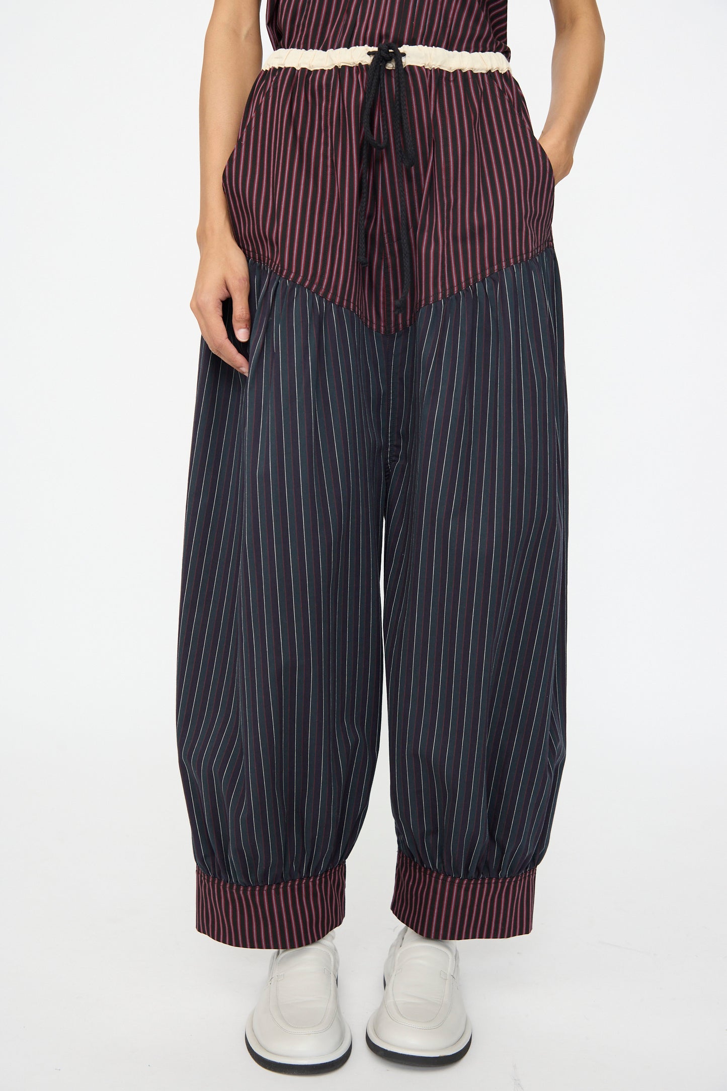 A person wears SC103’s Cotton Suiting Crescent Pants in Celestine Stripe, featuring loose, wide legs with maroon cuffs and waistband, paired with a matching top, a white drawstring belt, and white shoes.