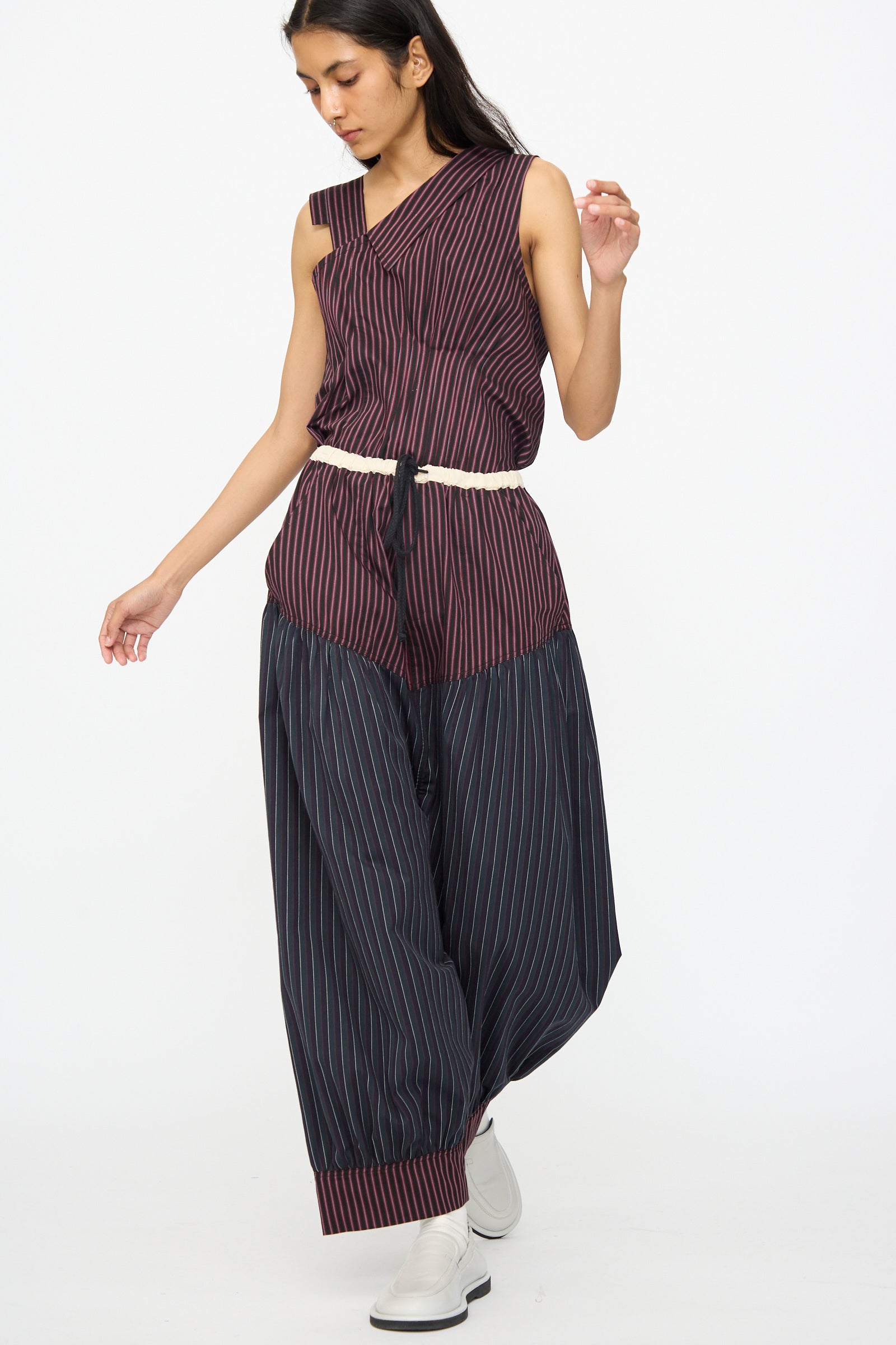A woman wearing SC103’s Cotton Suiting Crescent Pants in Celestine Stripe stands against a plain white background, styled with a sleeveless top, rope belt, and white shoes.