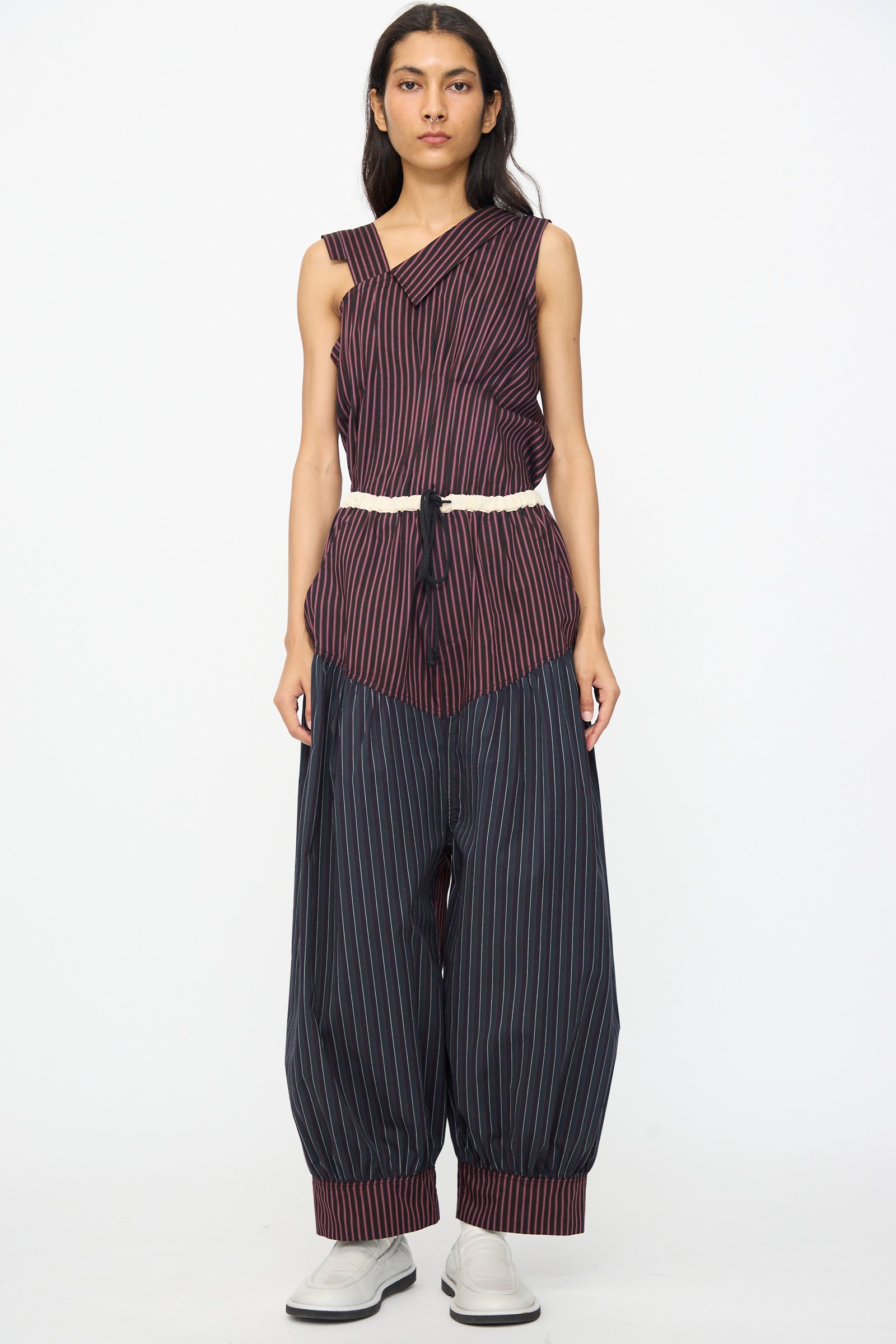 A person stands against a white background, wearing SC103 Cotton Suiting Crescent Pants in Celestine Stripe with a matching sleeveless asymmetrical striped top, paired with white shoes and a white belt.