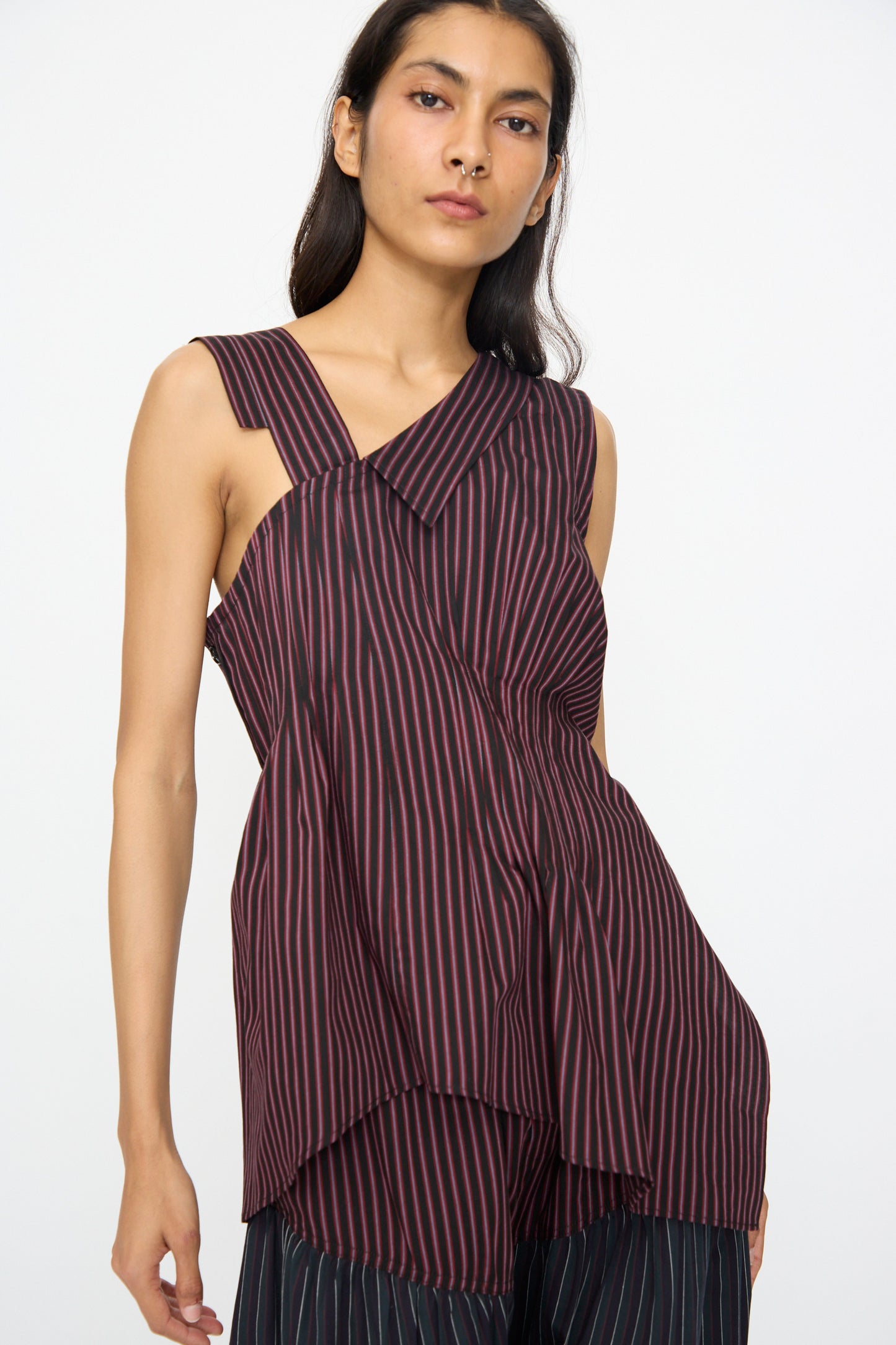 A person wearing the SC103 Cotton Suiting Solstice Top in Celestine Stripe—an asymmetrical, sleeveless piece with red and black vertical stripes—stands against a plain white background.