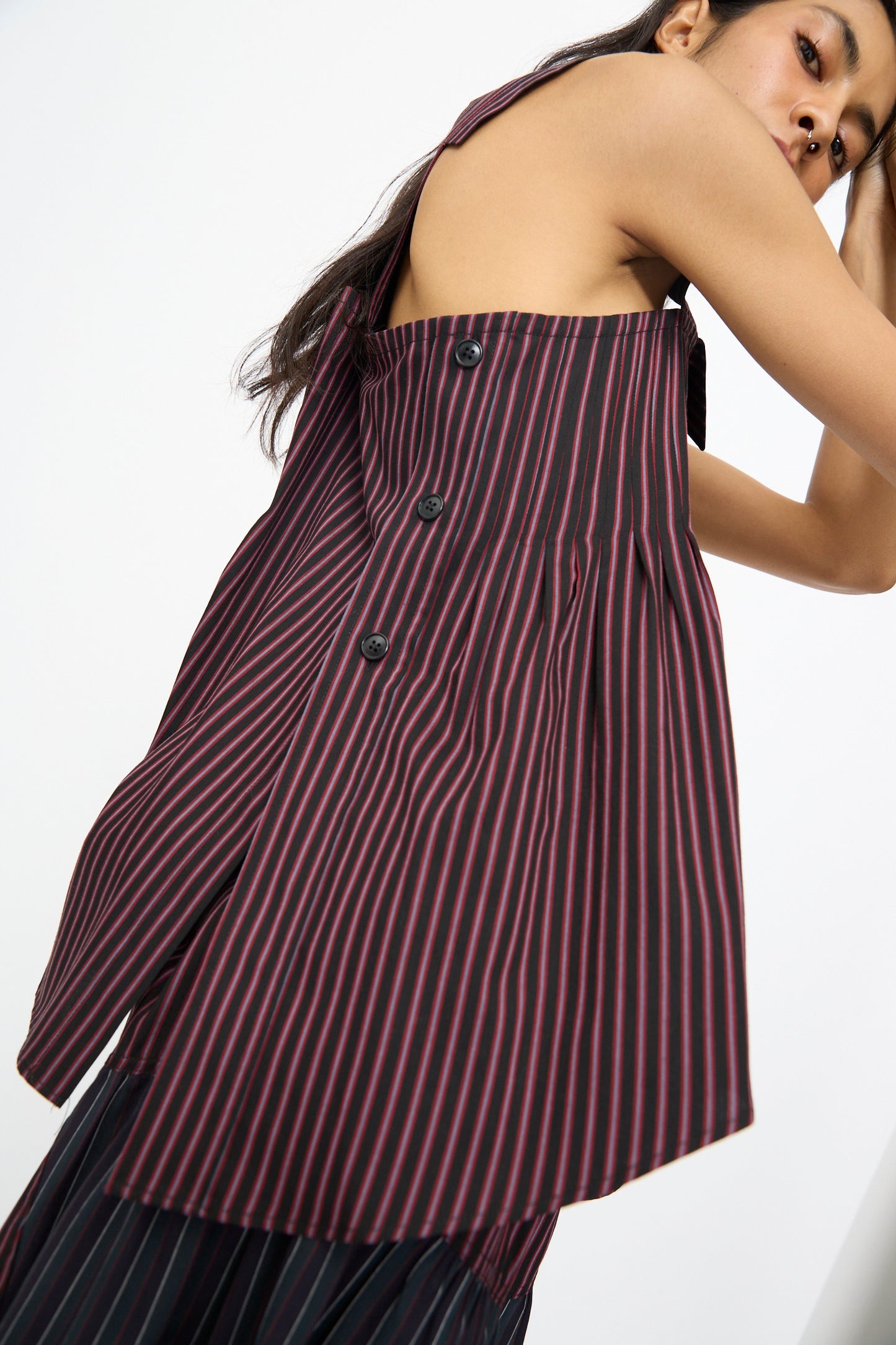 A woman models the SC103 Cotton Suiting Solstice Top in Celestine Stripe—an asymmetrical, sleeveless top with vertical maroon and black stripes and side buttons—posing with one arm raised near her face.