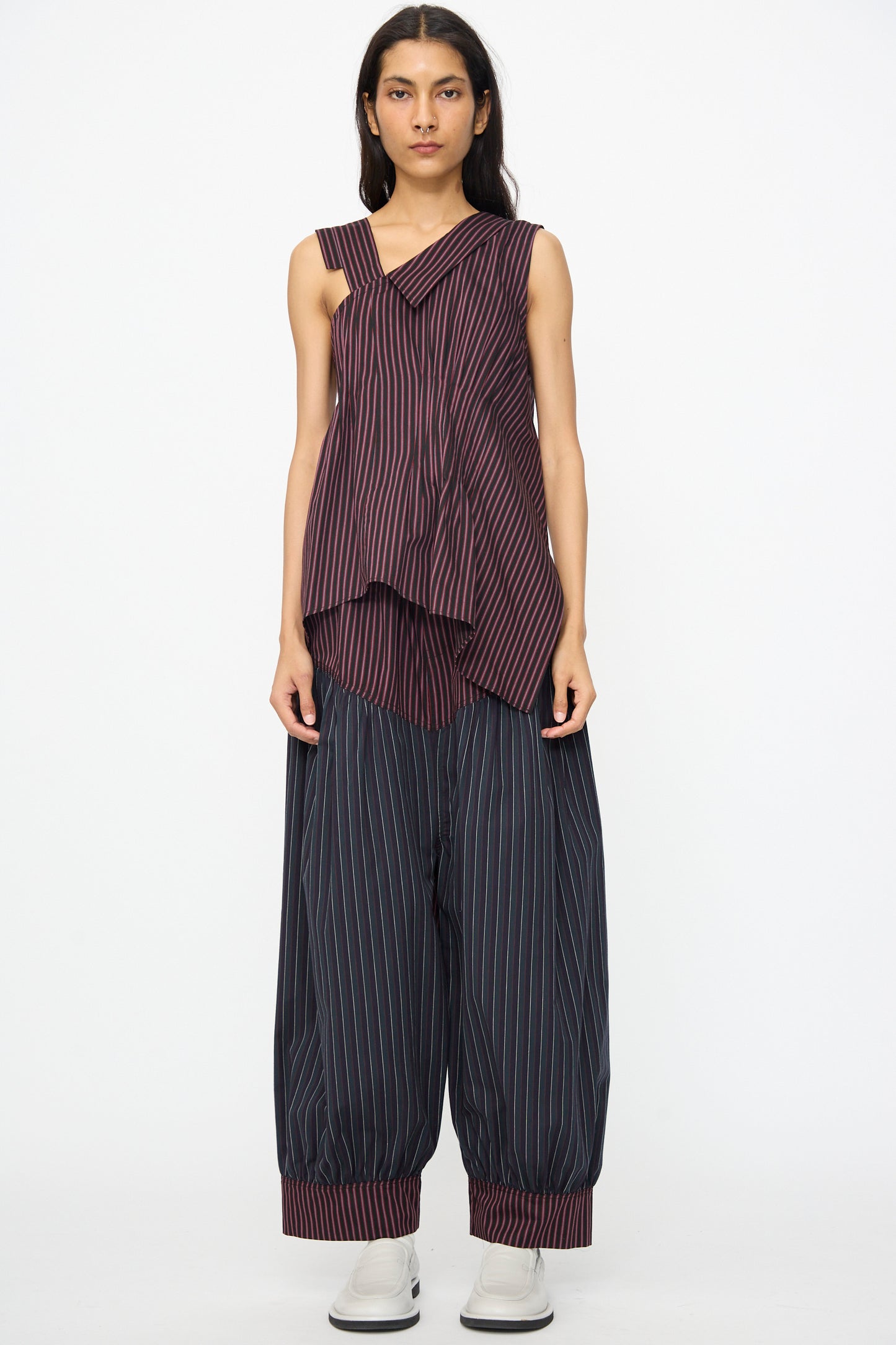 A person wears the SC103 Cotton Suiting Solstice Top in Celestine Stripe, an asymmetrical sleeveless striped cotton piece, paired with wide, cuffed striped pants and white shoes, standing against a plain white background. Made in NYC.