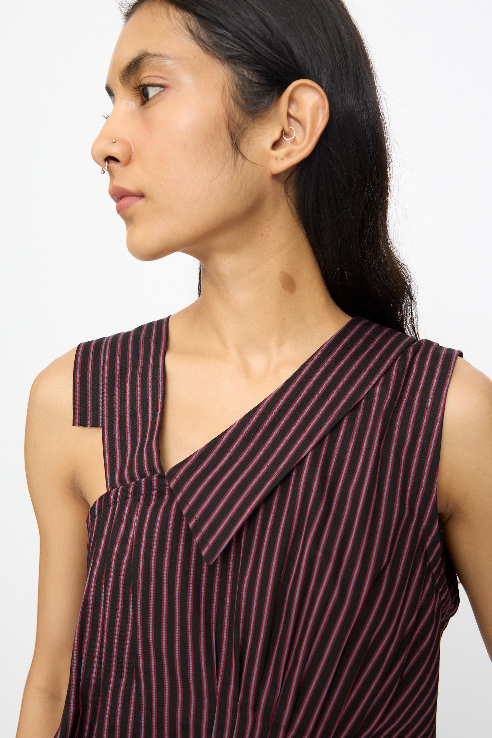 A person with long dark hair wears the SC103 Cotton Suiting Solstice Top in Celestine Stripe—an asymmetrical, sleeveless black and red pinstriped cotton top made in NYC—while looking left against a plain background.