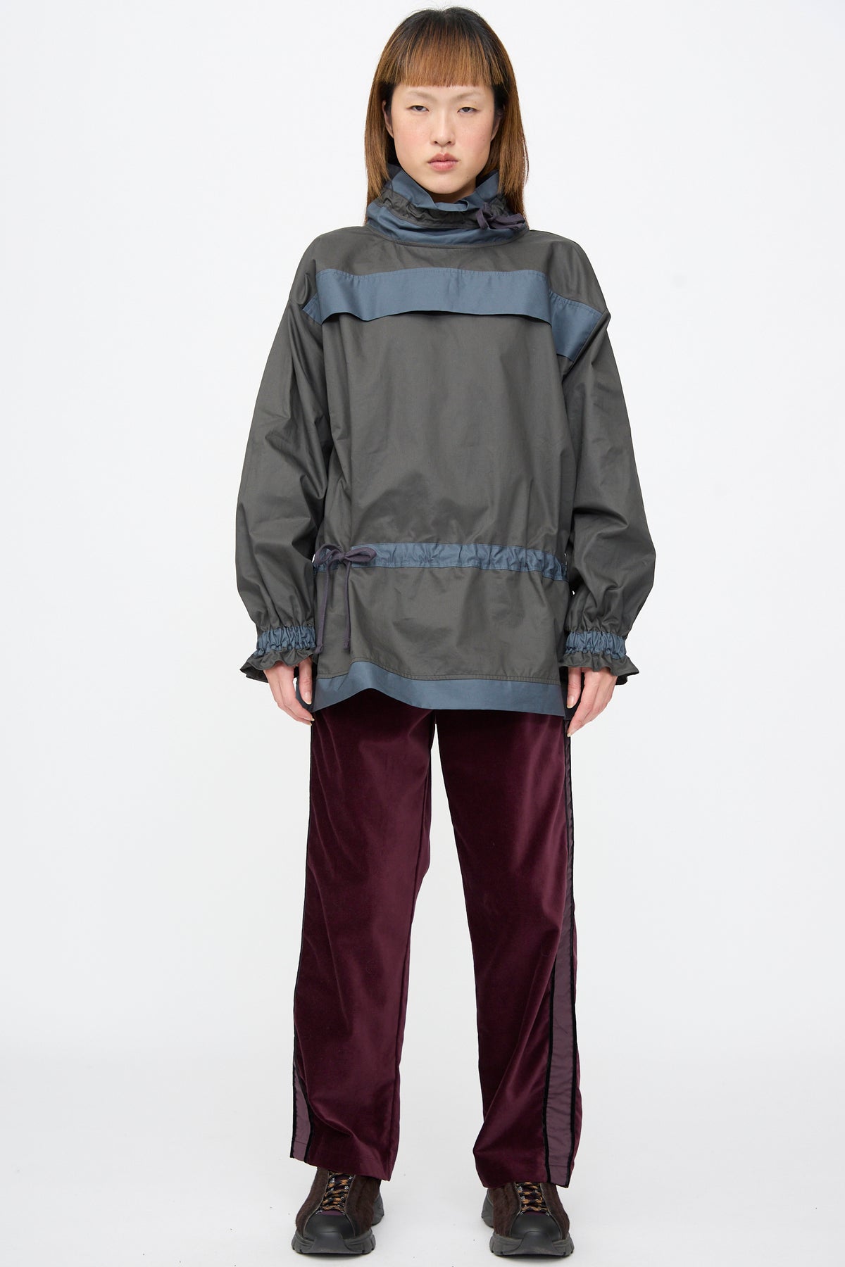 A person wears a dark grey windbreaker with a high collar over deep burgundy Cotton Velvet Chord Pants with side stripes and dark sneakers.