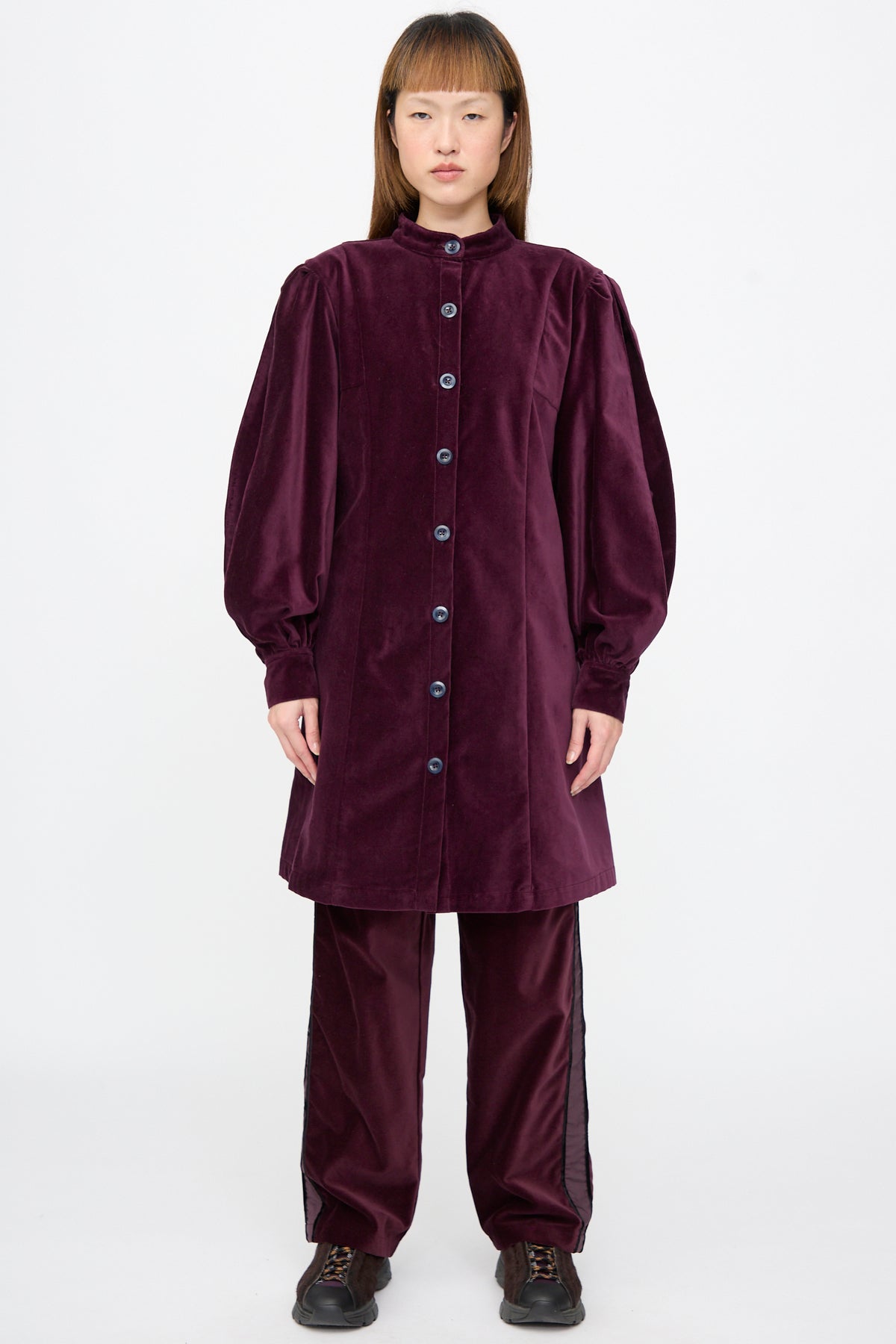A person wearing a SC103 Cotton Velvet Index Dress in Noble, a deep purple cotton velvet mini dress with balloon sleeves and matching trousers with a side stripe.