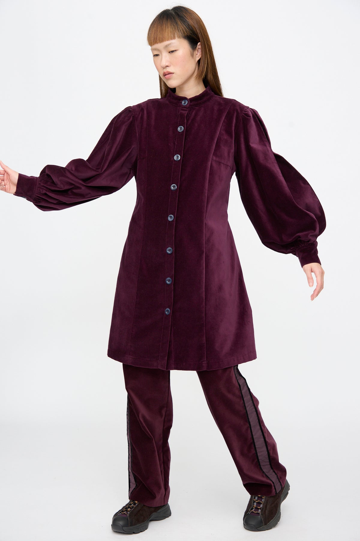 A person wears a deep purple SC103 Cotton Velvet Index Dress in Noble featuring balloon sleeves and button-front detailing, paired with matching trousers with side stripes and dark sneakers.