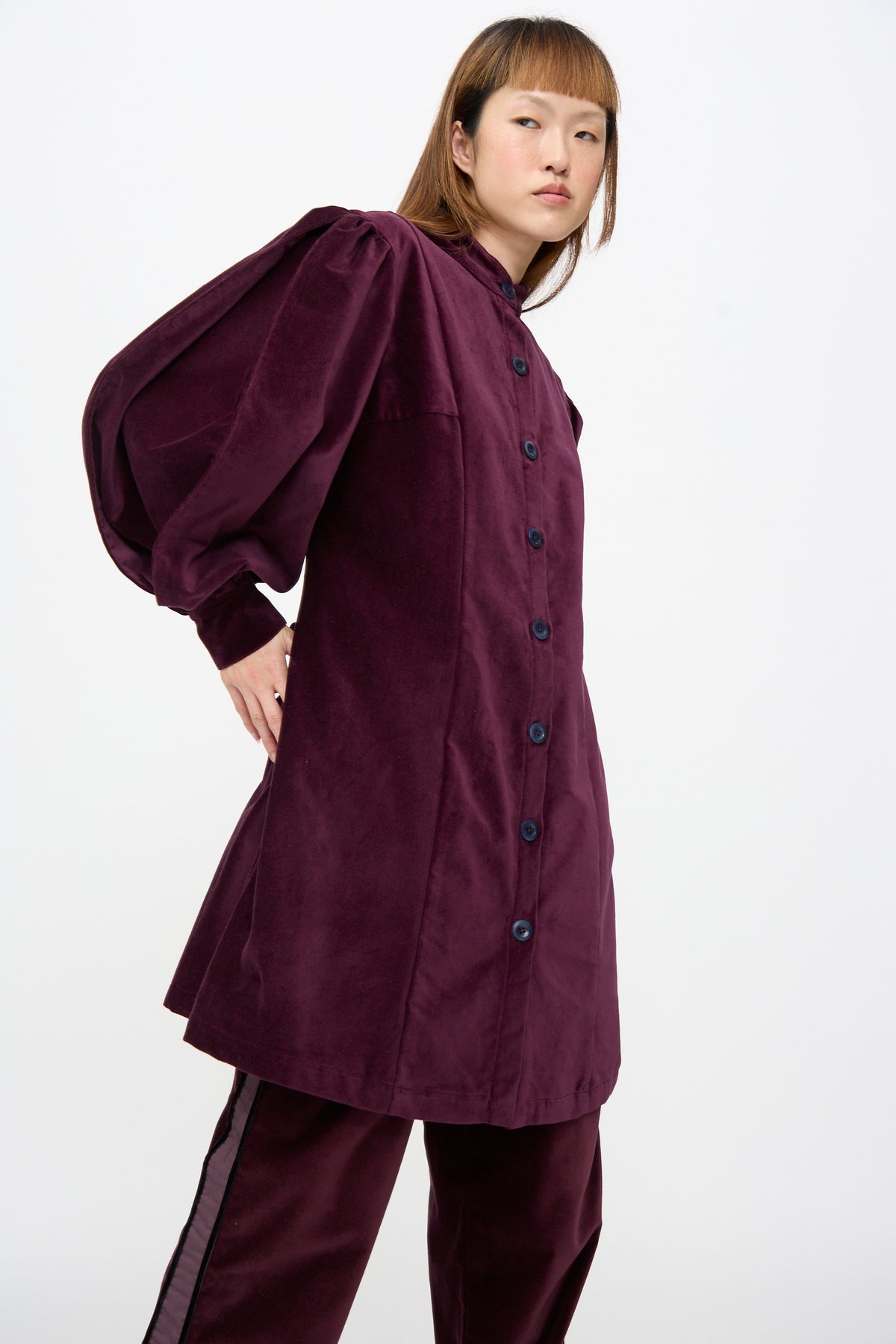 A person wearing a SC103 Cotton Velvet Index Dress in Noble, a deep purple cotton velvet mini dress with balloon sleeves and matching trousers with a side stripe.