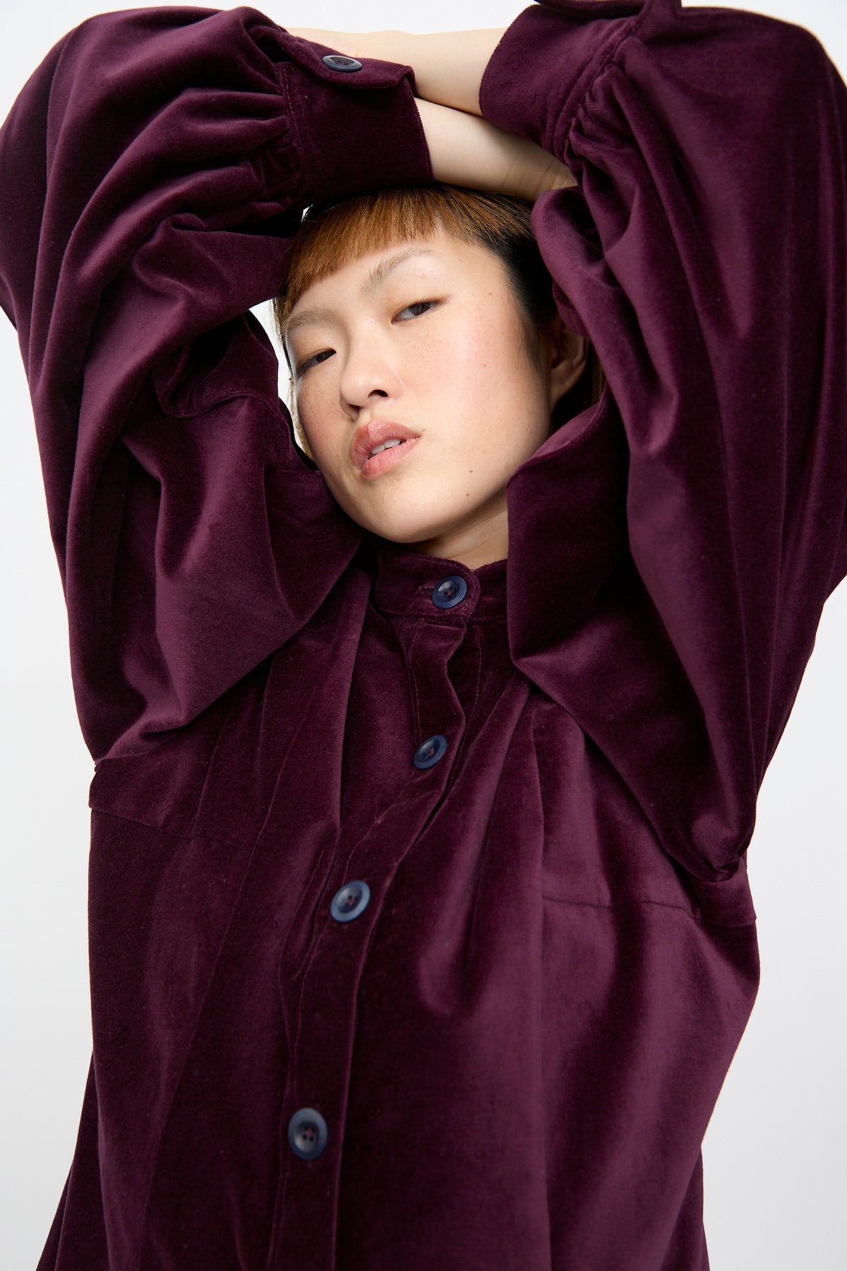 A person posing with arms raised behind their head, wearing the SC103 Cotton Velvet Index Dress in Noble, a plum-colored cotton velvet mini dress with balloon sleeves.