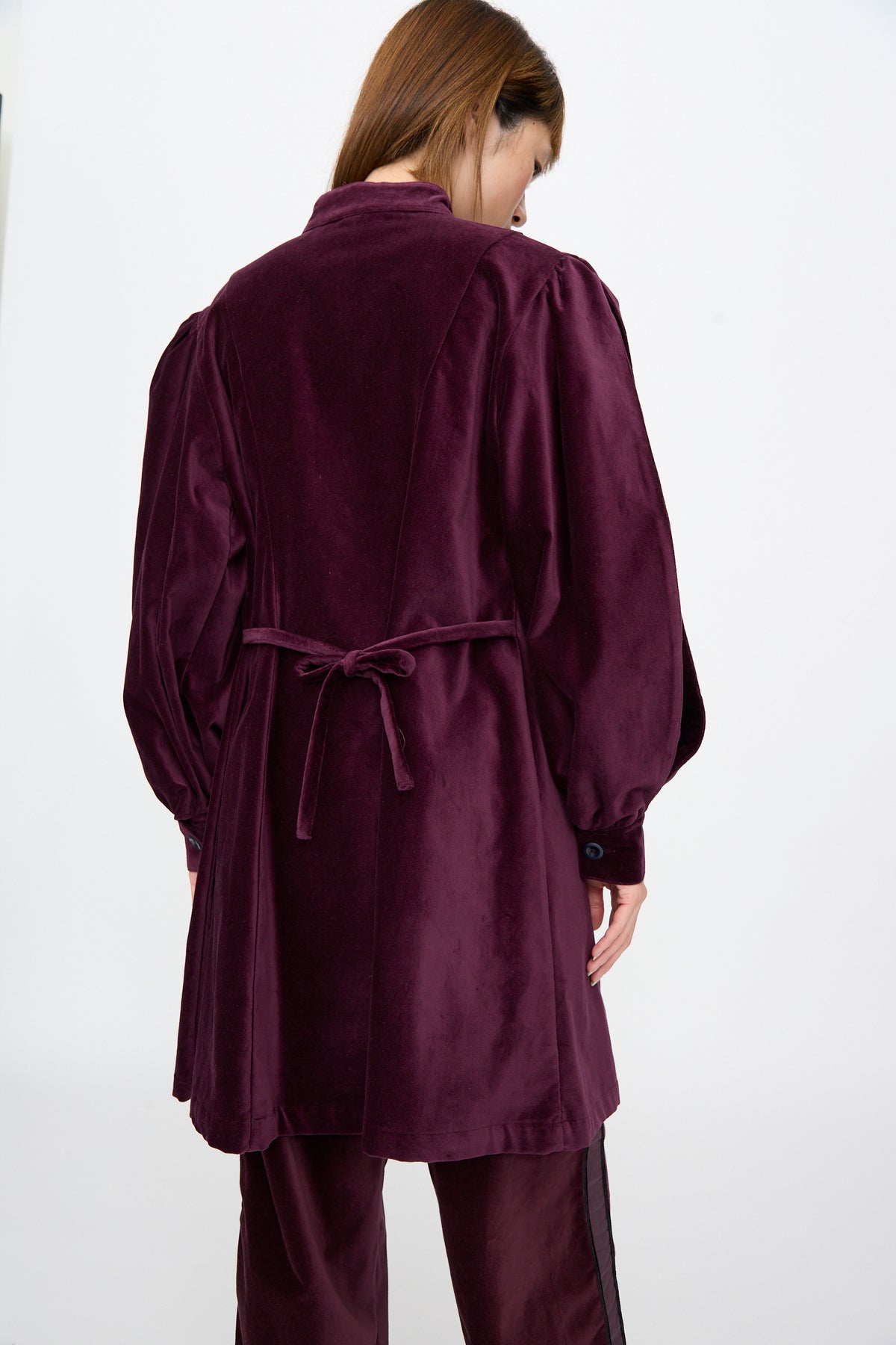 A person viewed from behind wearing a deep purple Cotton Velvet Index Dress in Noble featuring a tie-waist and balloon sleeves.