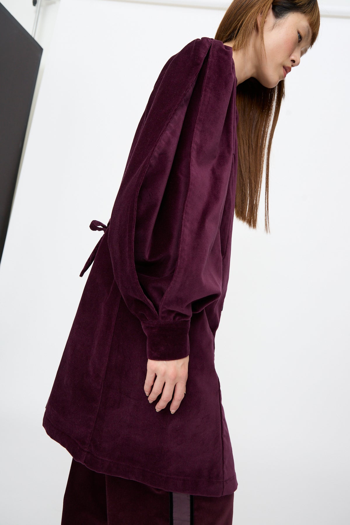 A person wearing a dark purple SC103 Cotton Velvet Index Dress in Noble, a long-sleeved mini dress with balloon sleeves and a tie at the waist.