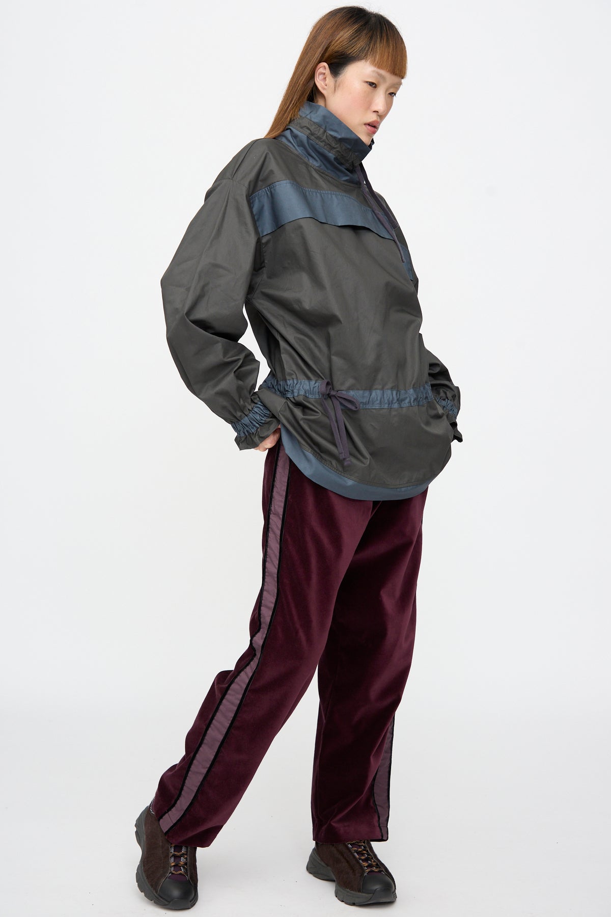 A person wears a dark-colored Crisp Cotton Incline Tunic in Twilight with a high neck and drawstring waist, paired with deep red trousers with side stripes and dark sneakers.