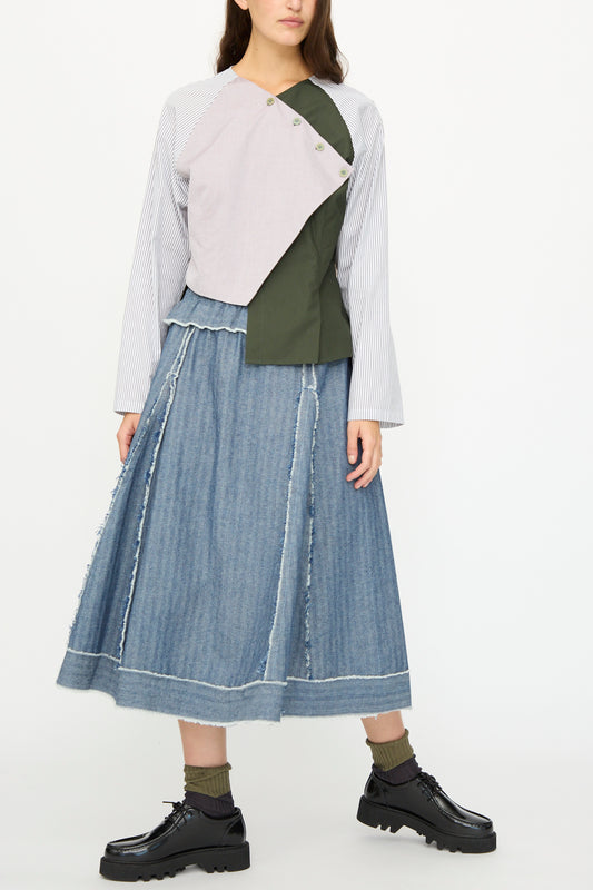 A woman wears SC103's Denim Radio Skirt in Herringbone, paired with black shoes, green socks, and a long-sleeve asymmetrical colorblock top in white, lilac, and olive green.