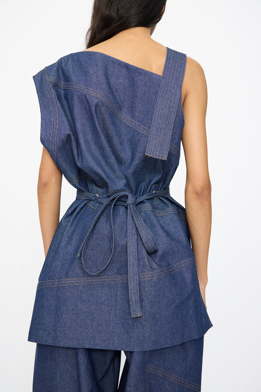 A woman is seen from the back wearing the SC103 Denim Sail Top in Soft Indigo, featuring a sleeveless design, asymmetrical neckline, and self-tie belt at the waist.