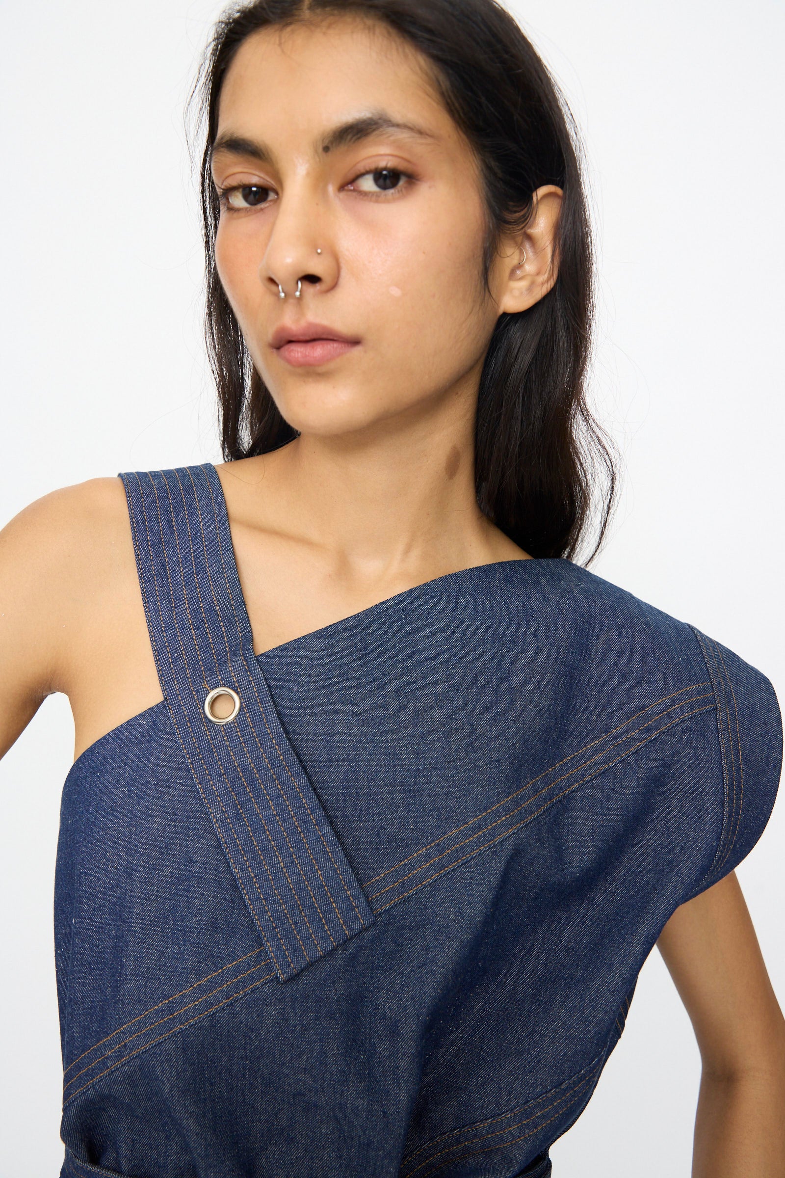 A person with long dark hair and a nose ring wears the SC103 Denim Sail Top in Soft Indigo, which features an asymmetrical neckline and visible stitching, standing against a plain background.