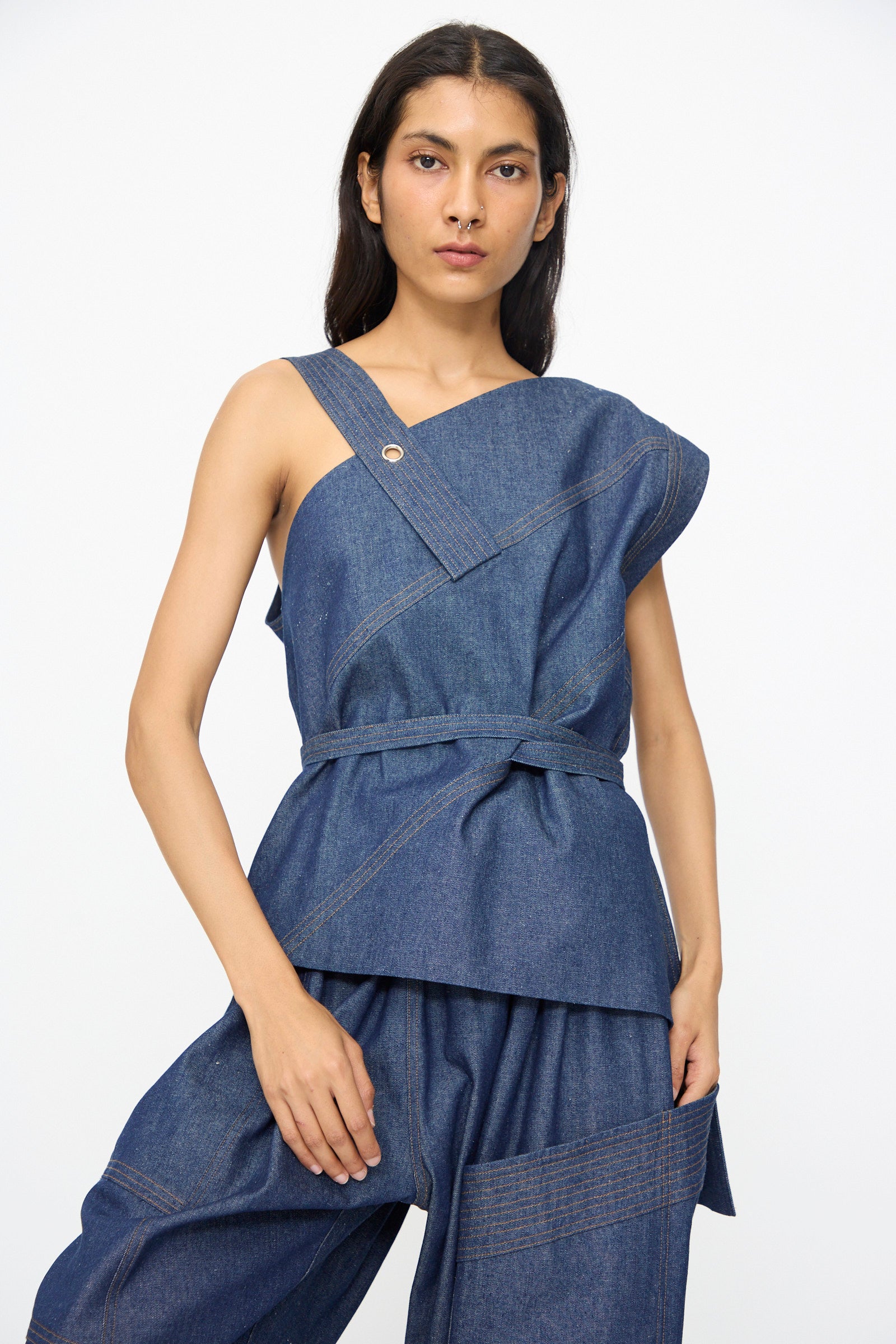 A person wears the SC103 Denim Sail Top in Soft Indigo—sleeveless with an asymmetrical neckline and self-tie belt—paired with matching wide-leg pants, standing against a plain white background.