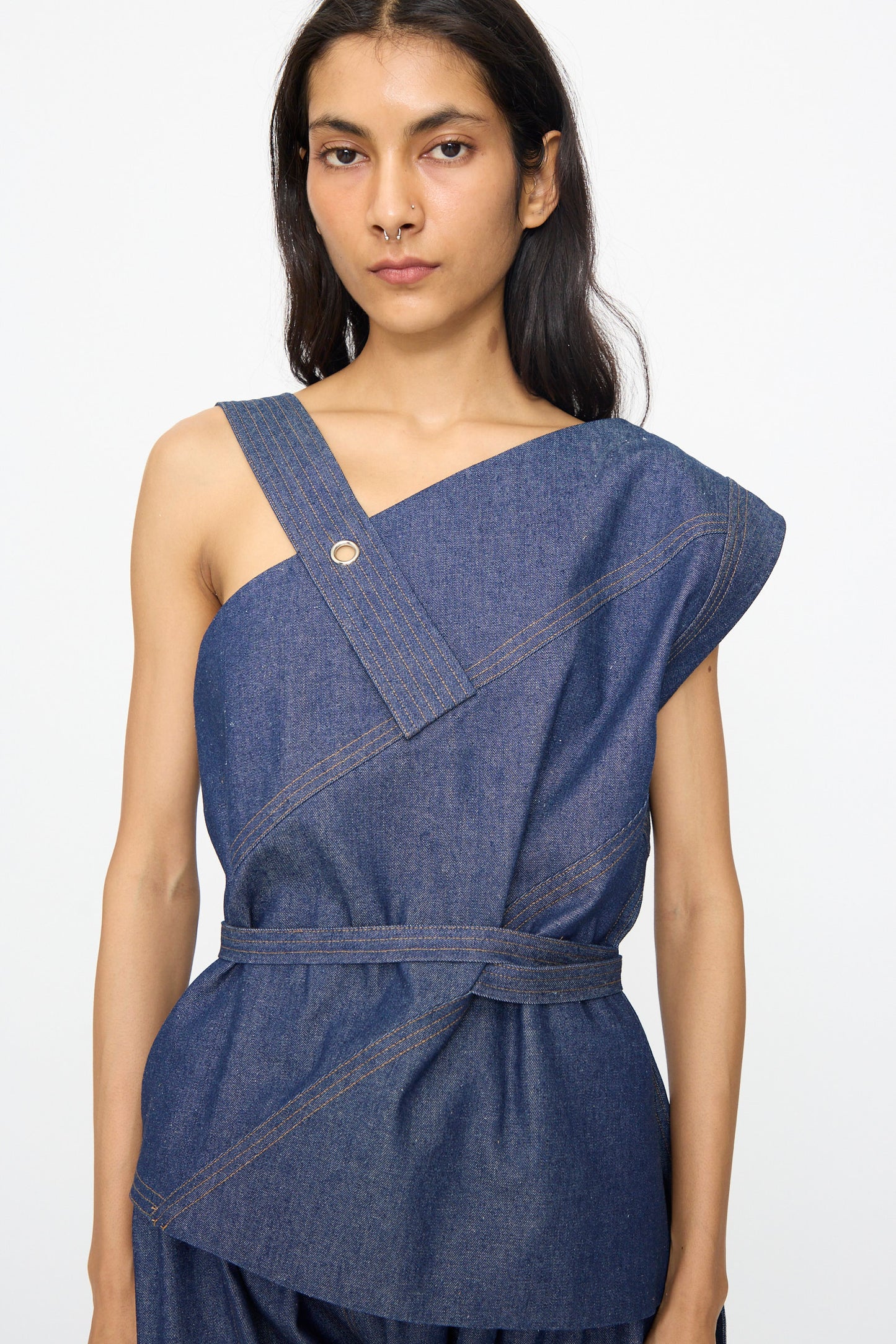 A person models the SC103 Denim Sail Top in Soft Indigo, featuring a sleeveless design, asymmetrical neckline, self-tie belt, and button details, standing against a plain background.