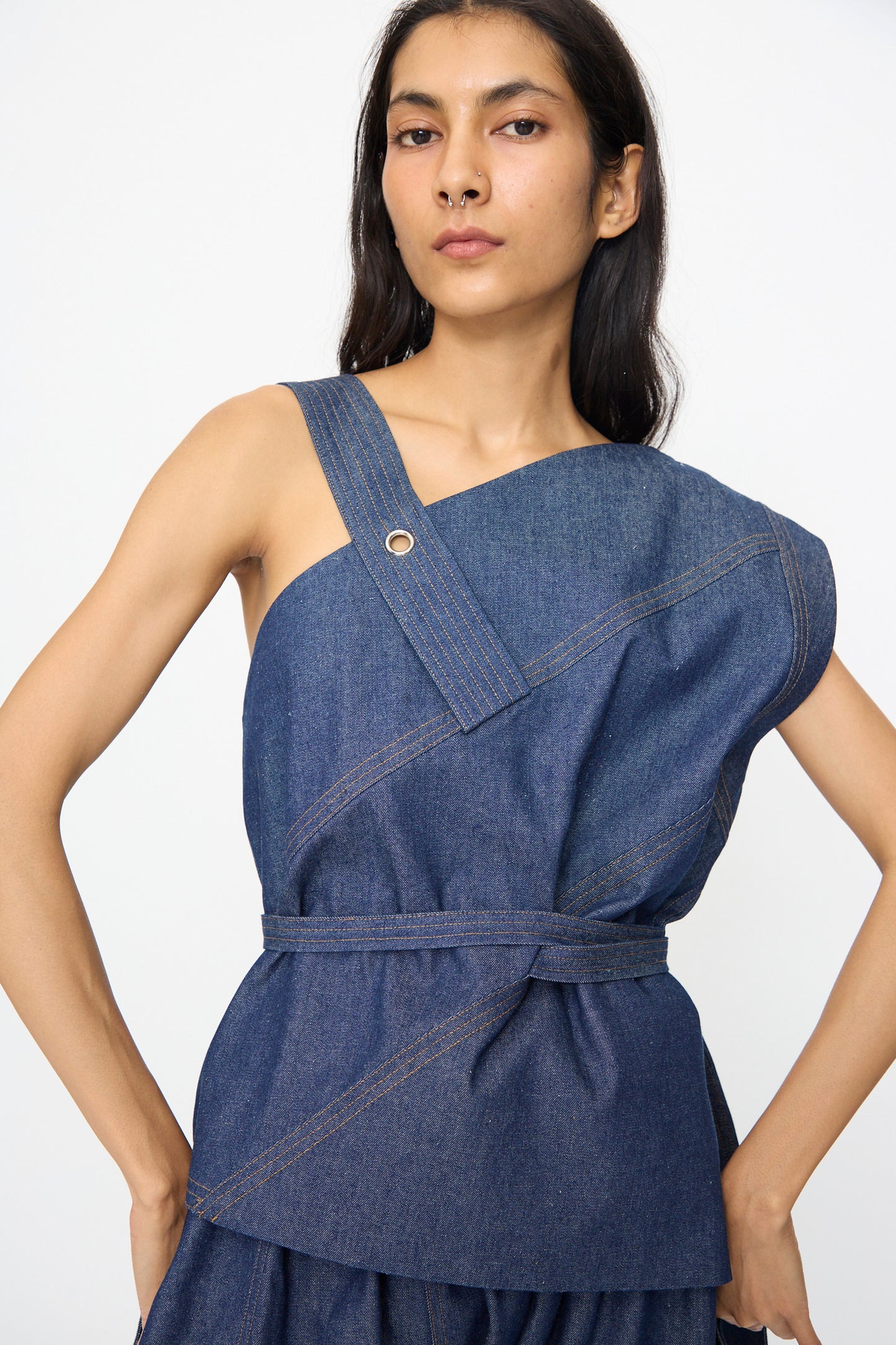 A person wears the SC103 Denim Sail Top in Soft Indigo, a sleeveless piece with an asymmetrical neckline, self-tie belt, and visible stitching, standing against a plain background.