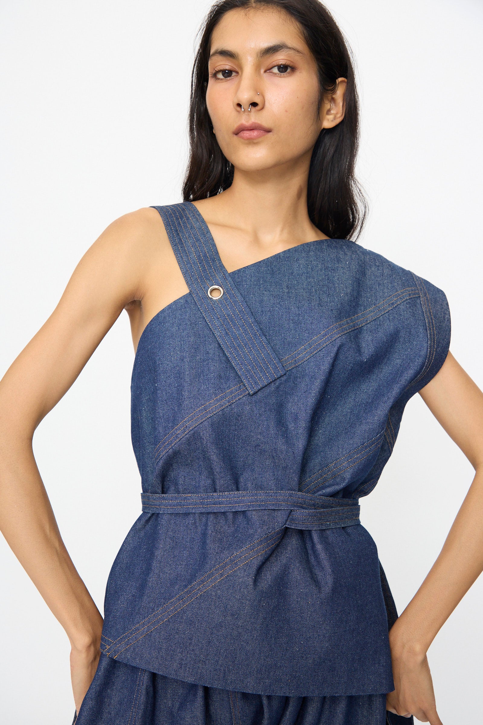 A person wears the SC103 Denim Sail Top in Soft Indigo, a sleeveless piece with an asymmetrical neckline, self-tie belt, and visible stitching, standing against a plain background.