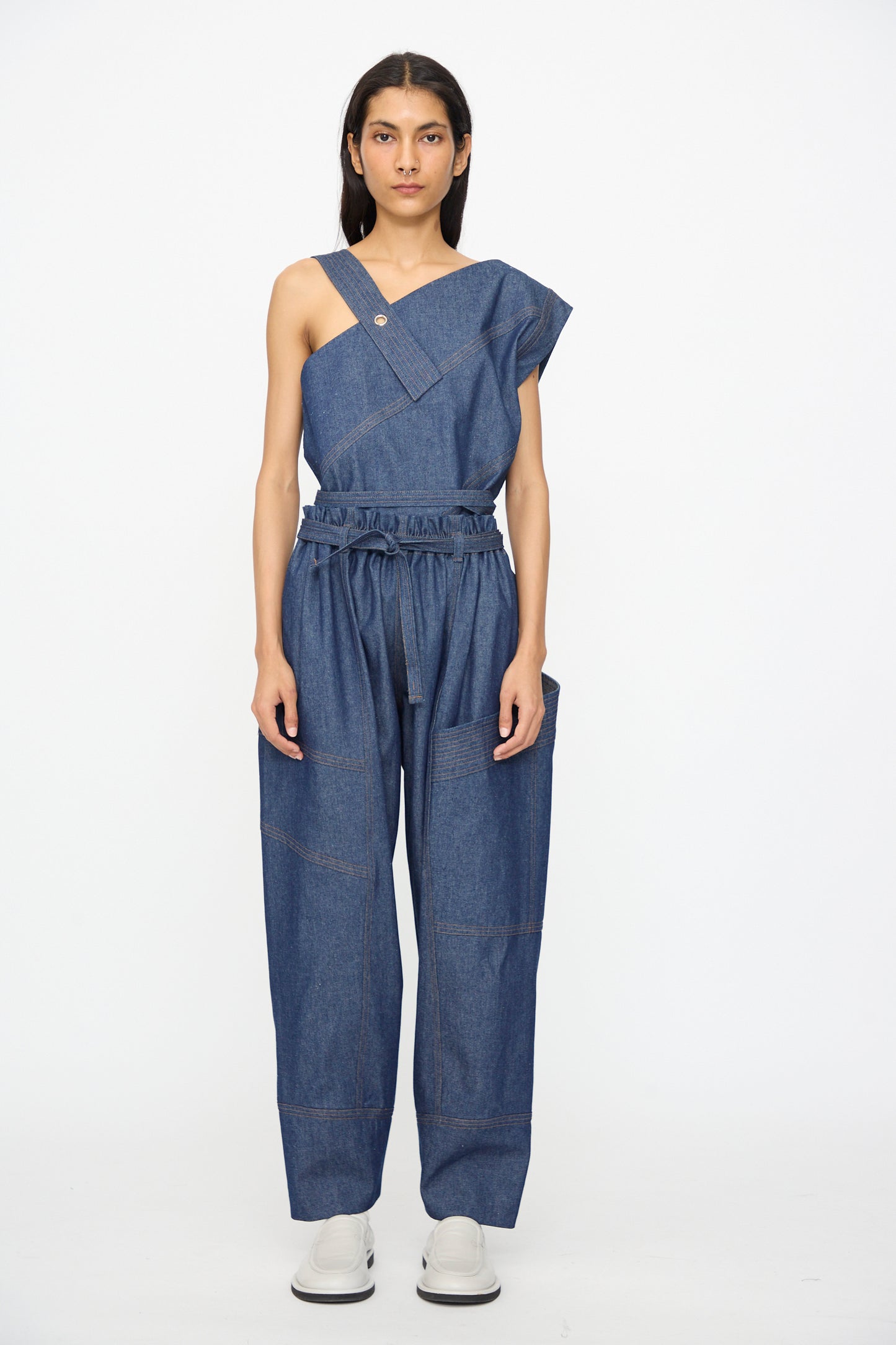 A person stands against a plain background wearing an asymmetrical, one-shoulder denim top and SC103 Denim Vault Pants in Soft Indigo with a drawstring waist, paired with crisp white shoes.