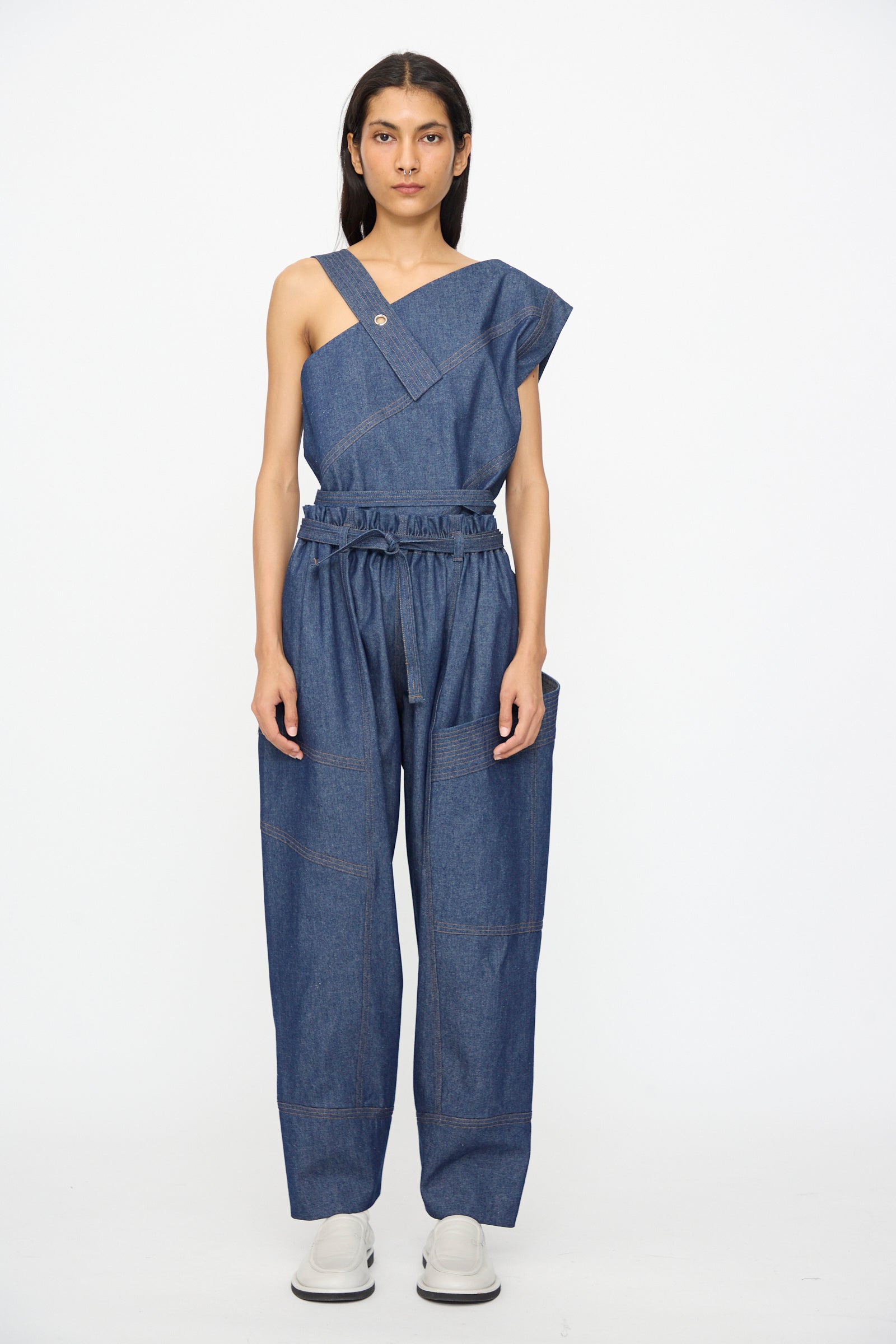 A person stands against a plain background wearing an asymmetrical, one-shoulder denim top and SC103 Denim Vault Pants in Soft Indigo with a drawstring waist, paired with crisp white shoes.