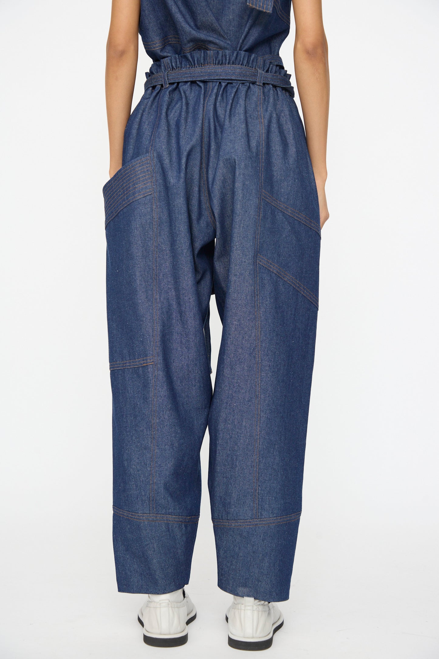 A person faces away, wearing SC103 Denim Vault Pants in Soft Indigo—relaxed fit, high-waisted with large pockets—and white shoes, set against a plain white background.