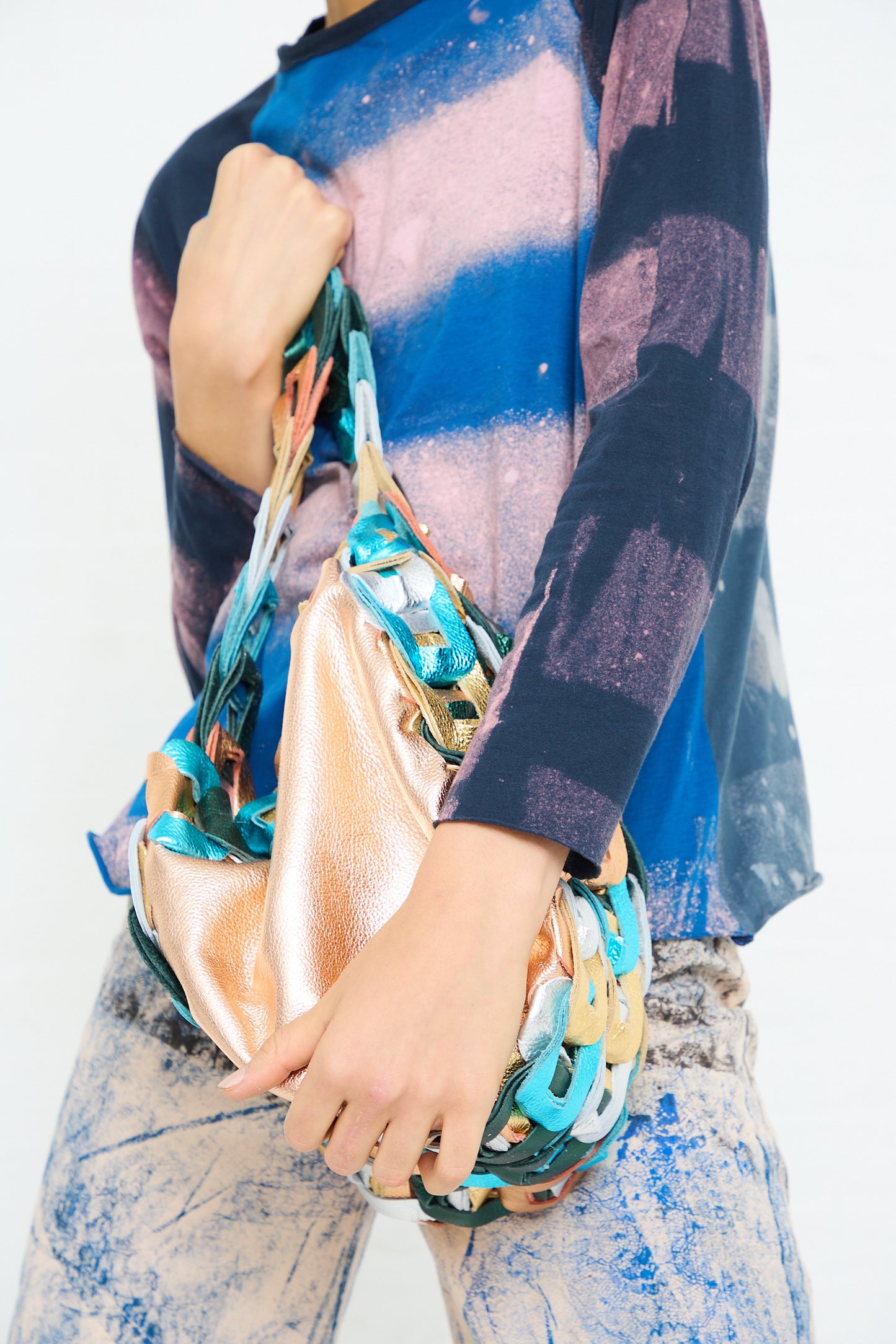 A person holding a shiny, multicolored SC103 Leather Drum Bag in Mineral with a leather links strap, against a backdrop of a cosmic-themed shirt and splattered jeans.