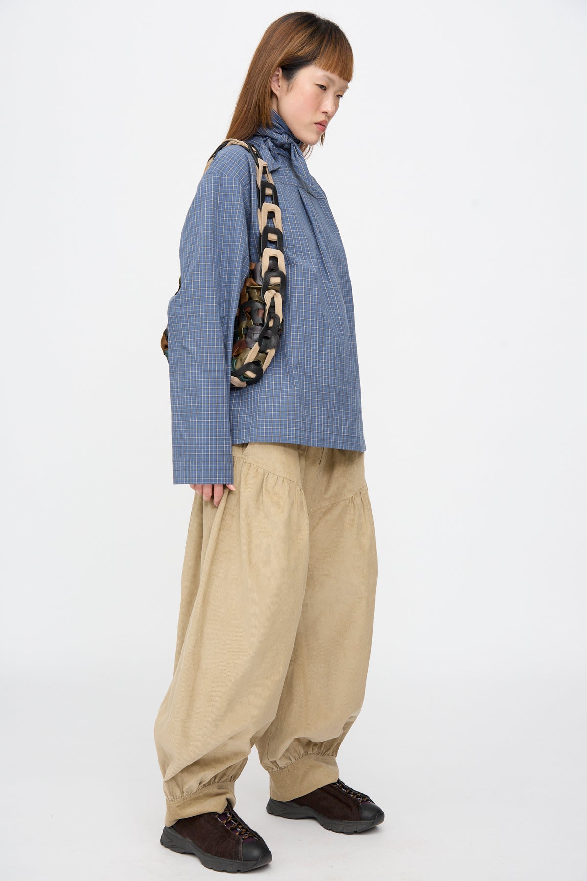 A person wearing a blue plaid shirt and tan trousers carries a SC103 Leather Links Tote in Fortress, featuring interlocking, multi-colored leather links.