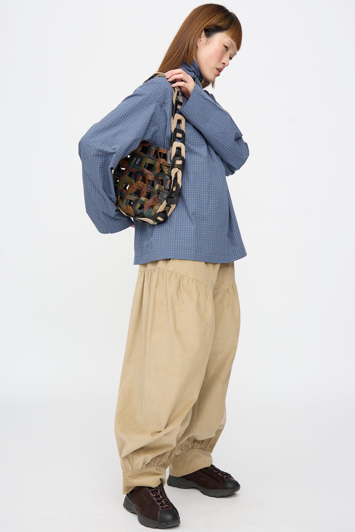 A person in a blue plaid shirt and tan pants carries a SC103 Leather Links Tote in Fortress, featuring interlocking multicolored leather links and wide strap.
