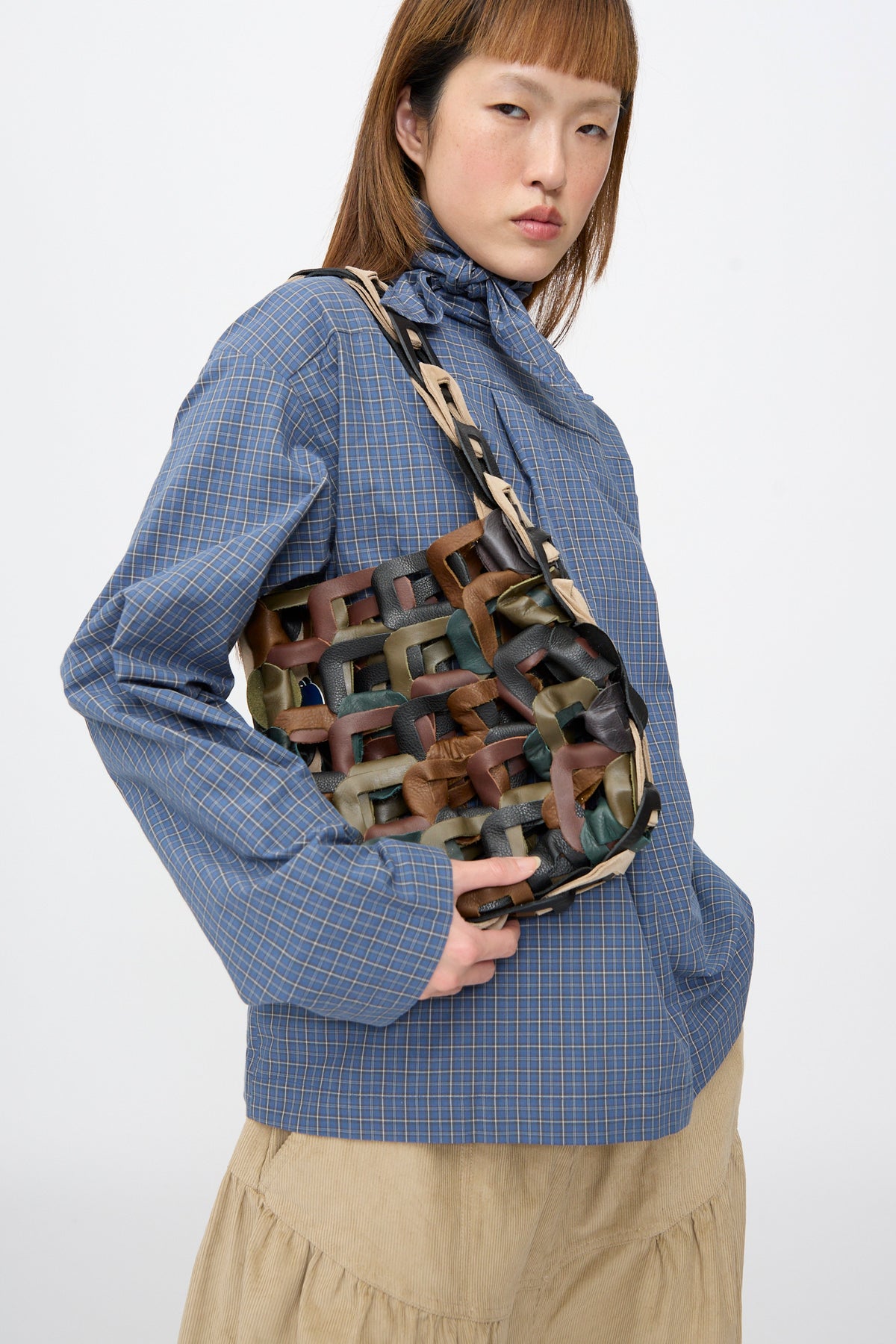 A person in a blue plaid shirt carries the SC103 Leather Links Tote in Fortress, featuring interlocking dark green, brown, and tan leather links.