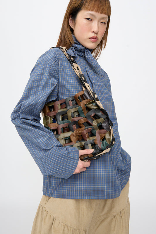 A person in a blue plaid shirt carries the SC103 Leather Links Tote in Fortress, featuring interlocking dark green, brown, and tan leather links.