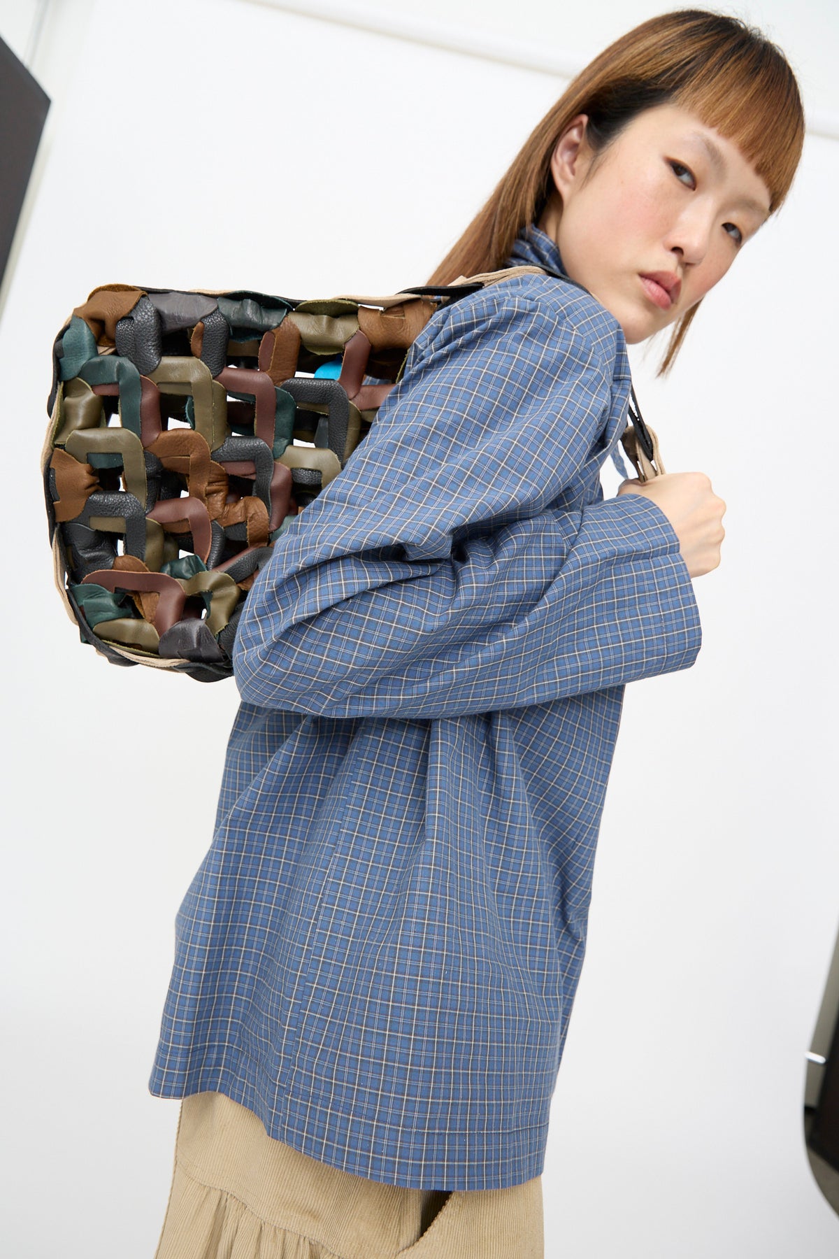 A person wears a blue plaid shirt and carries the SC103 Leather Links Tote in Fortress, featuring interlocking links in brown, green, and black leather